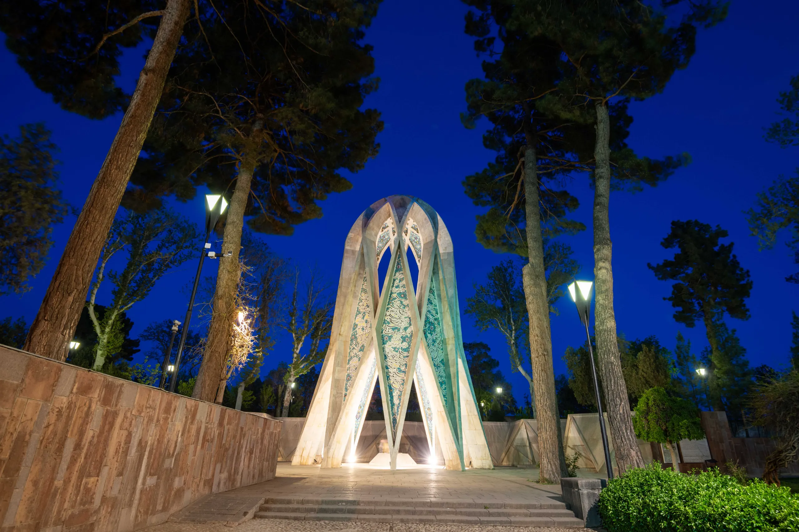 A modern, geometric mausoleum with turquoise patterns, illuminated at night under a dark blue sky, flanked by tall pine trees and streetlights. Photograph by Mohammad Ataei.