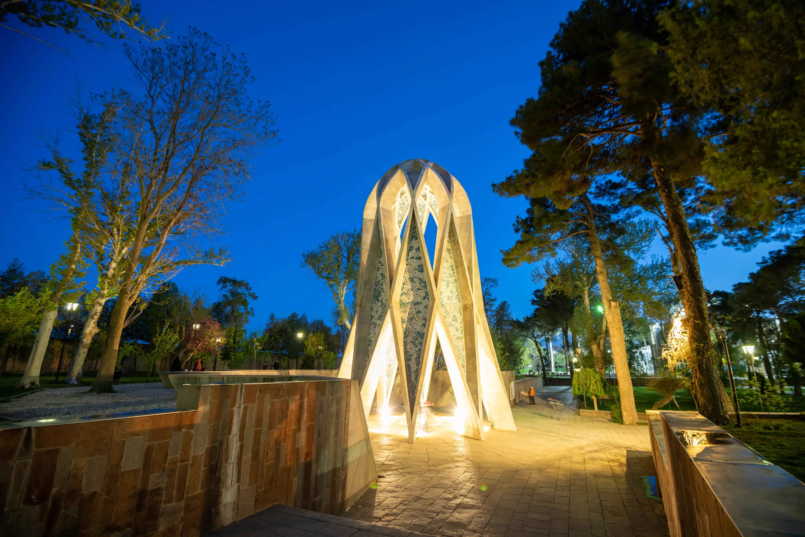 A strikingly illuminated, modern geometric mausoleum stands prominently at night amidst lush trees and paved walkways, under a deep blue sky. Photograph by Mohammad Ataei.
