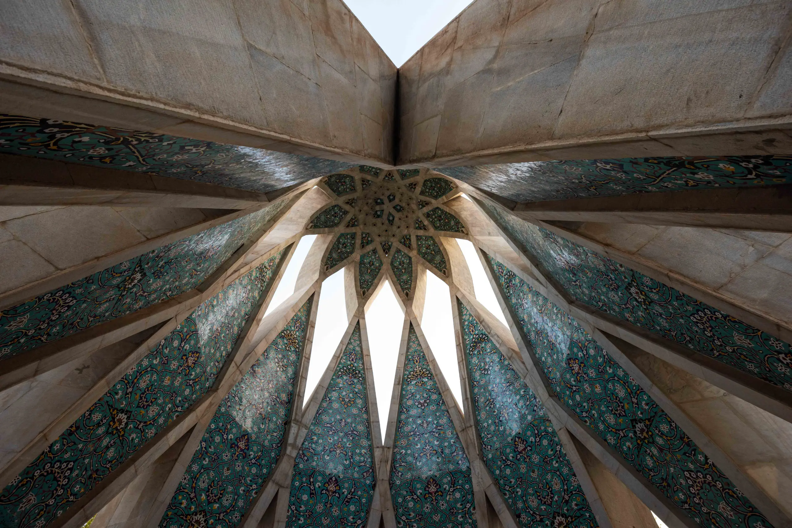 A low-angle shot looking up into the interior of a geometrically patterned dome or vault, with teal and white tiles, and open spaces letting in light. Photograph by Mohammad Ataei.