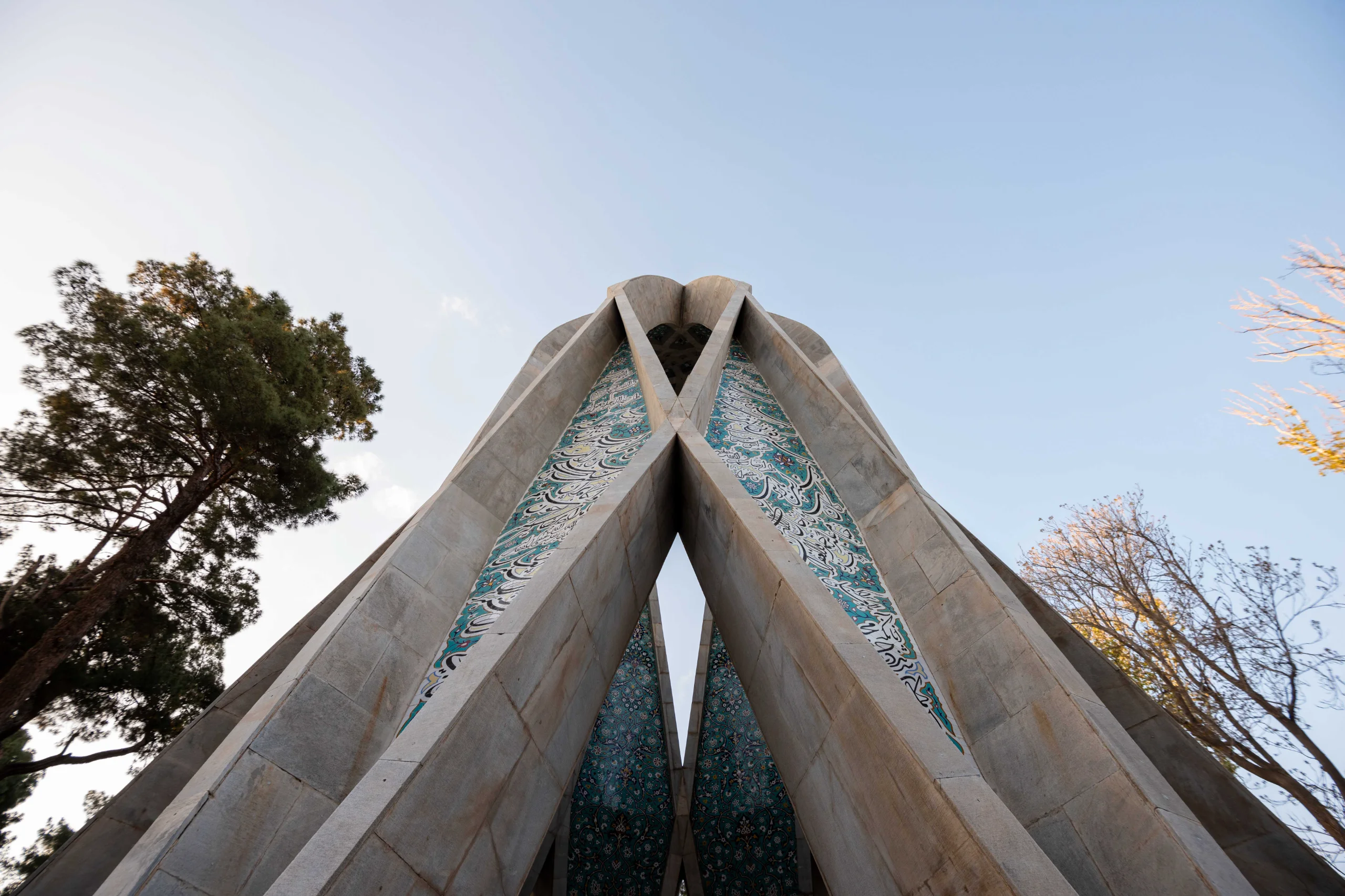 A low-angle shot of Omar Khayyam's Mausoleum shows its modern, geometric structure tapering upwards, adorned with intricate turquoise and white tilework. Tall trees flank the monument against a clear blue sky. Photograph by Mohammad Ataei.