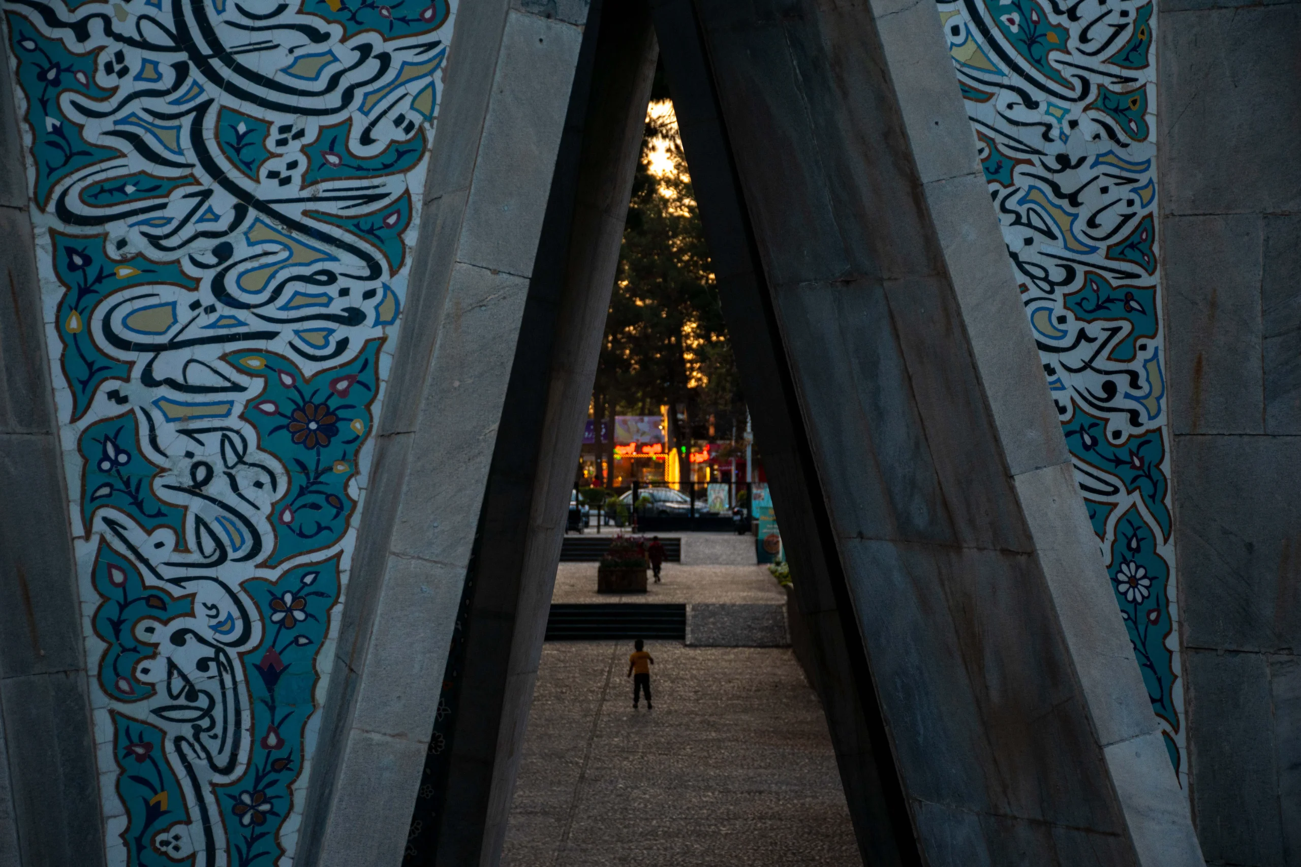 A framed perspective through the concrete pylons and turquoise tilework of Khayyam's mausoleum looking out toward a paved courtyard, with a distant figure walking. Photograph by Mohammad Ataei.