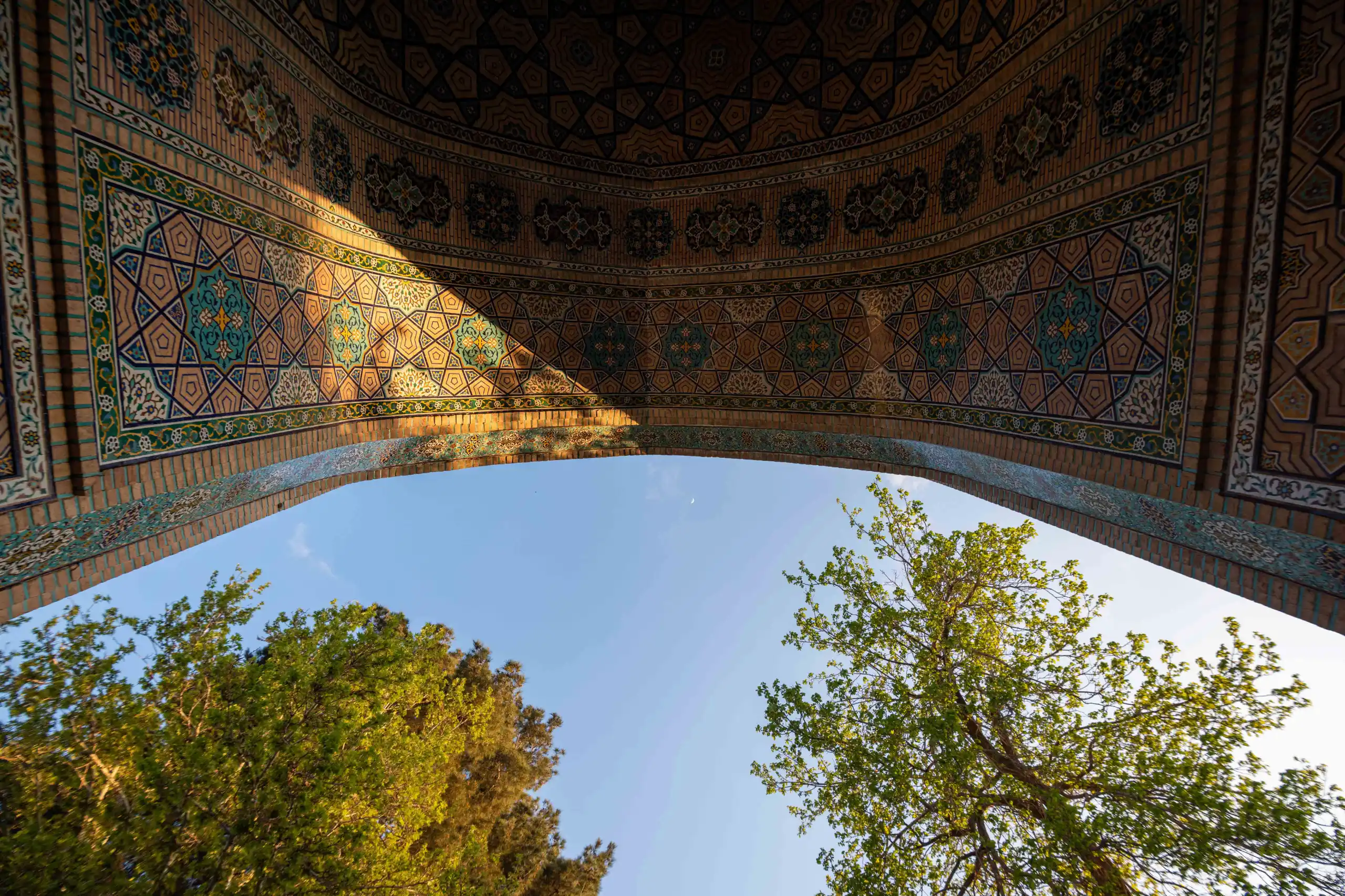 A low-angle shot shows a beautifully tiled archway with intricate geometric patterns, illuminated by a diagonal beam of sunlight, framing a clear blue sky and green tree branches. Photograph by Mohammad Ataei.