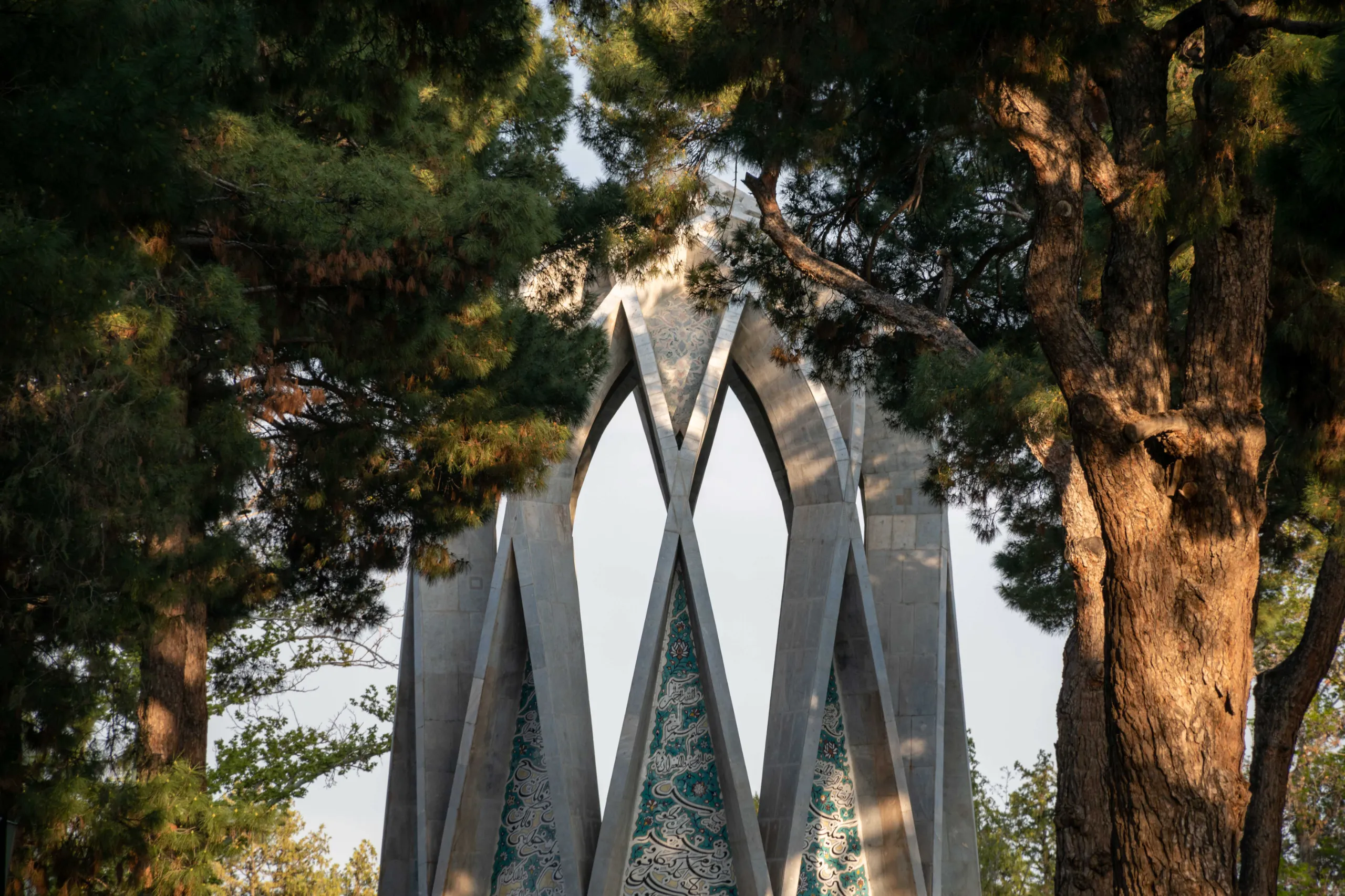The pointed arches and tiled pillars of the Khayyam Mausoleum in Nishapur framed by dense green pine trees under natural daylight. Photograph by Mohammad Ataei.