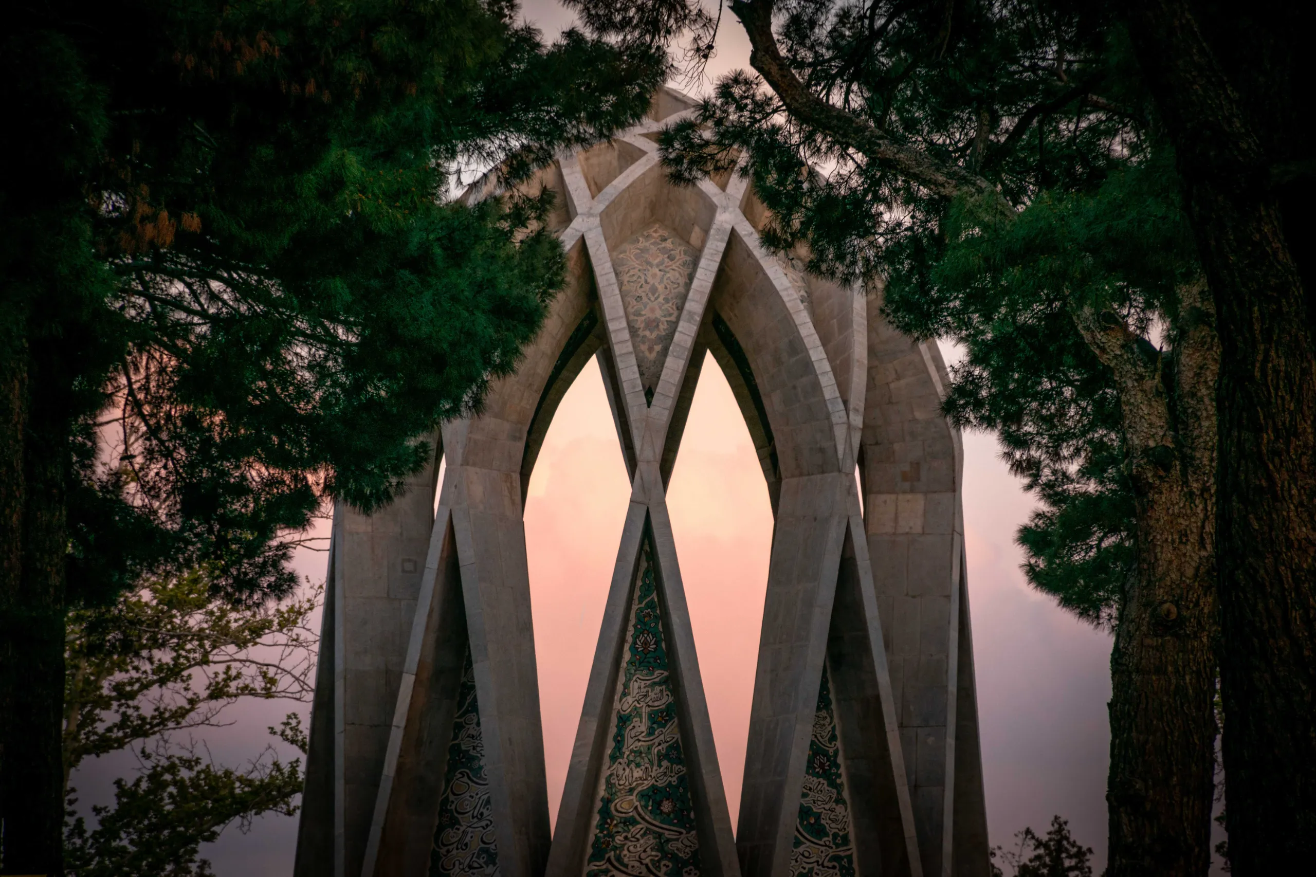 Looking through framing pine trees at the geometric concrete arches of the Khayyam Mausoleum against a pale sky. Photograph by Mohammad Ataei.