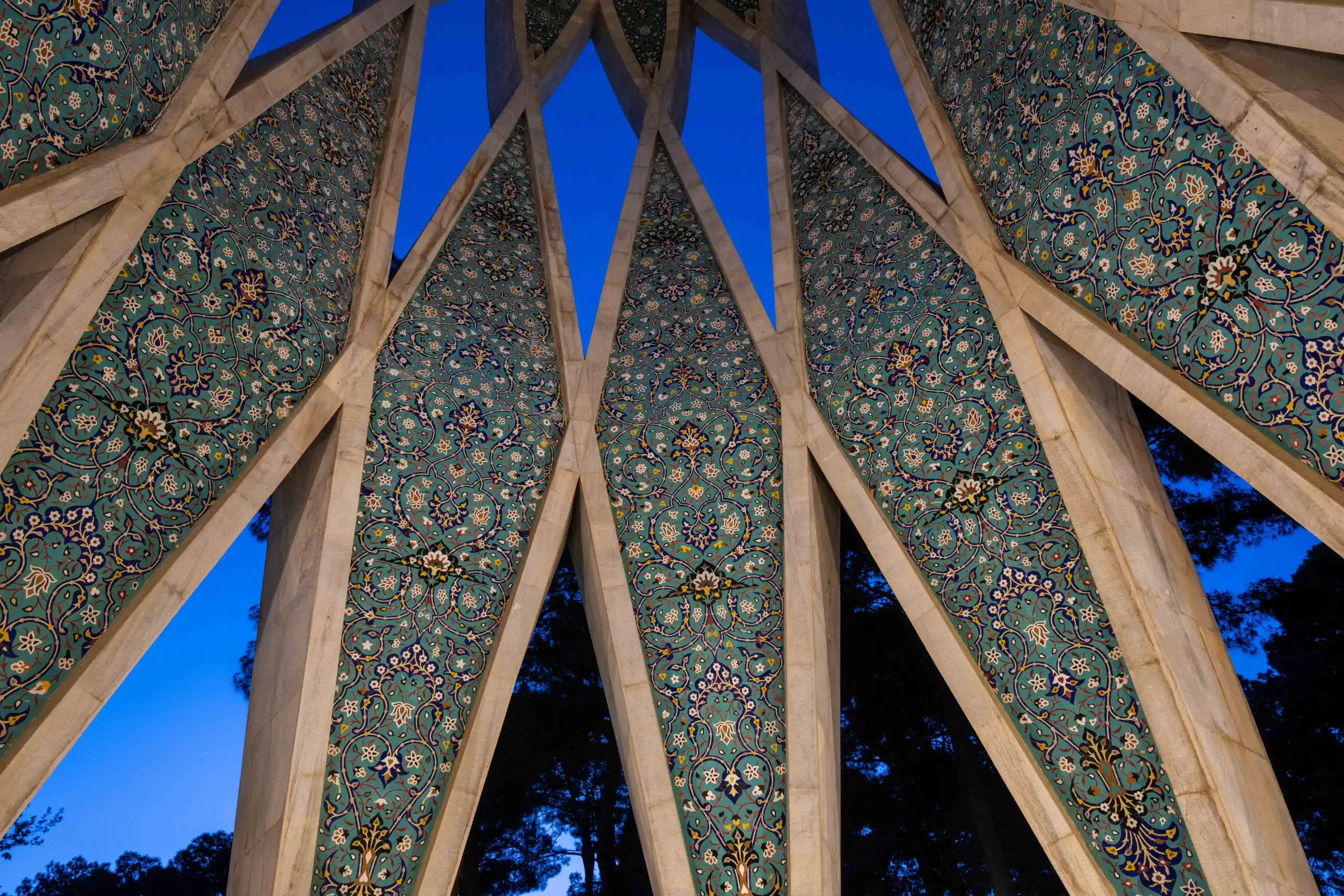 A low-angle view shows the geometrically intricate ceiling of the Khayyam Mausoleum, featuring repetitive turquoise and blue floral tile patterns against a dark blue evening sky. Photograph by Mohammad Ataei.