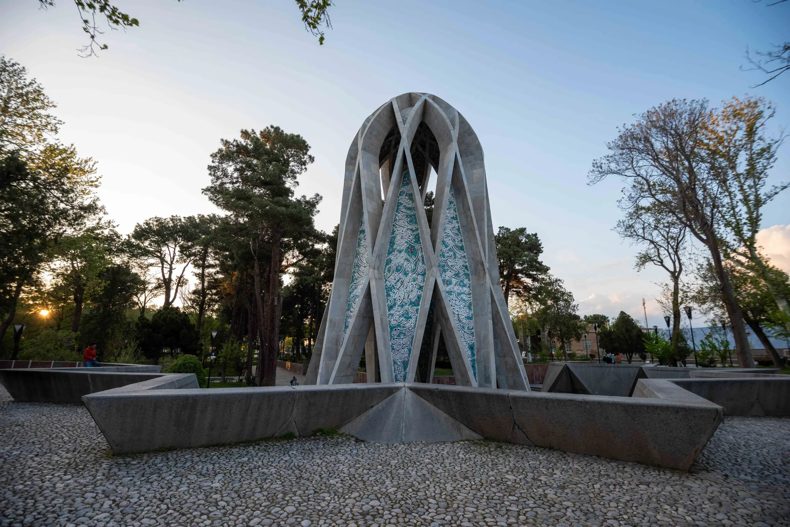 A striking modern mausoleum with a conical, ribbed concrete structure and turquoise tilework, surrounded by green trees and a pebble-paved ground under a clear sky at sunset. Photograph by Mohammad Ataei.
