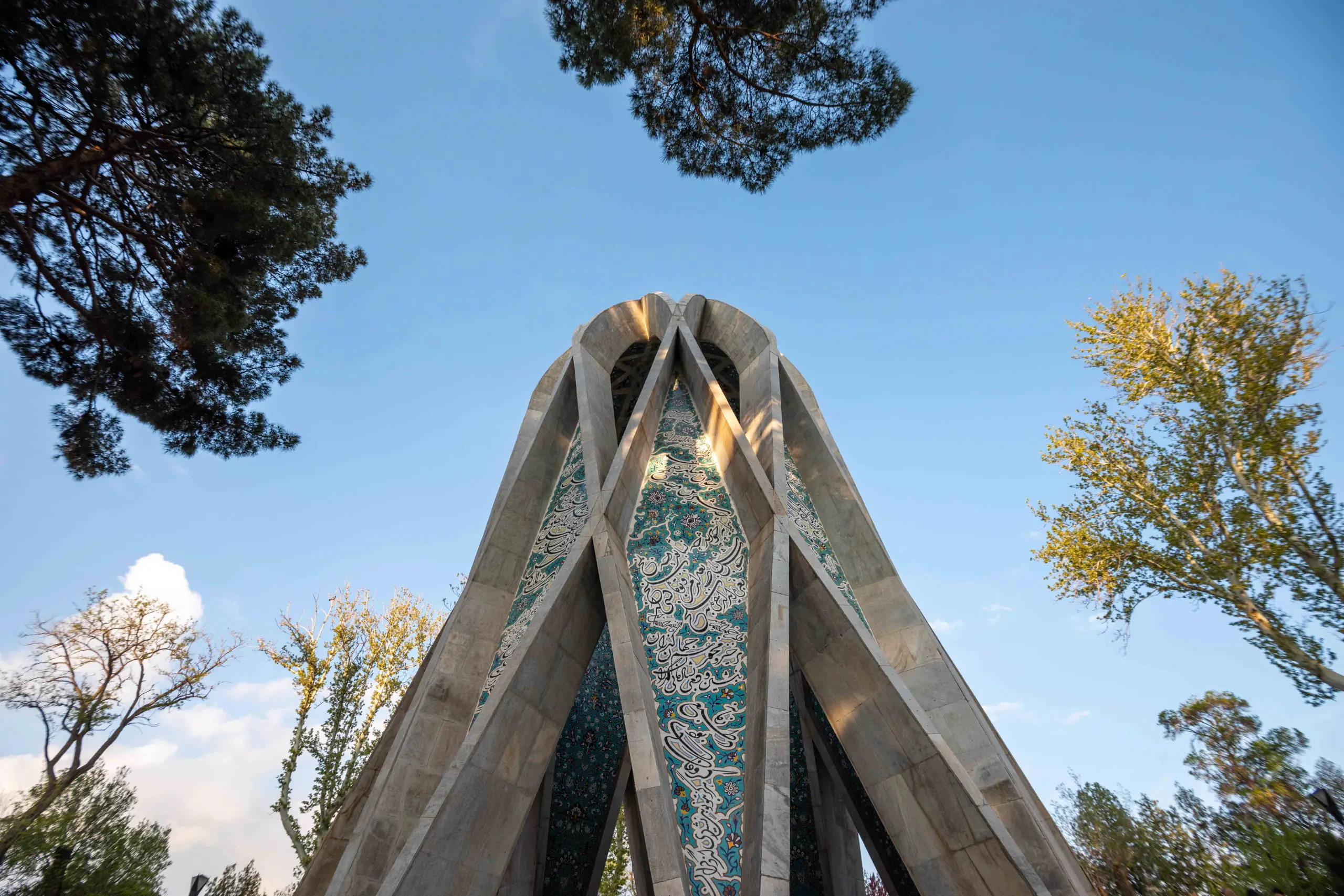 A low-angle view of a tall, modern, star-shaped mausoleum with geometric concrete-like elements and intricate turquoise and white tilework featuring Persian calligraphy, set against a bright blue sky and framed by green trees. Photograph by Mohammad Ataei.