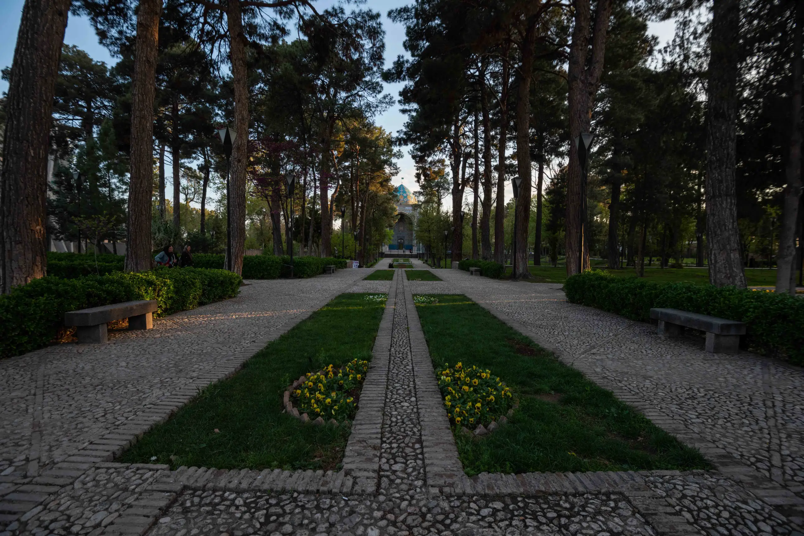 A long, symmetrical garden path lined with tall trees and green lawns leads to a dome-topped mausoleum in the distance under a clear sky. Photograph by Mohammad Ataei.