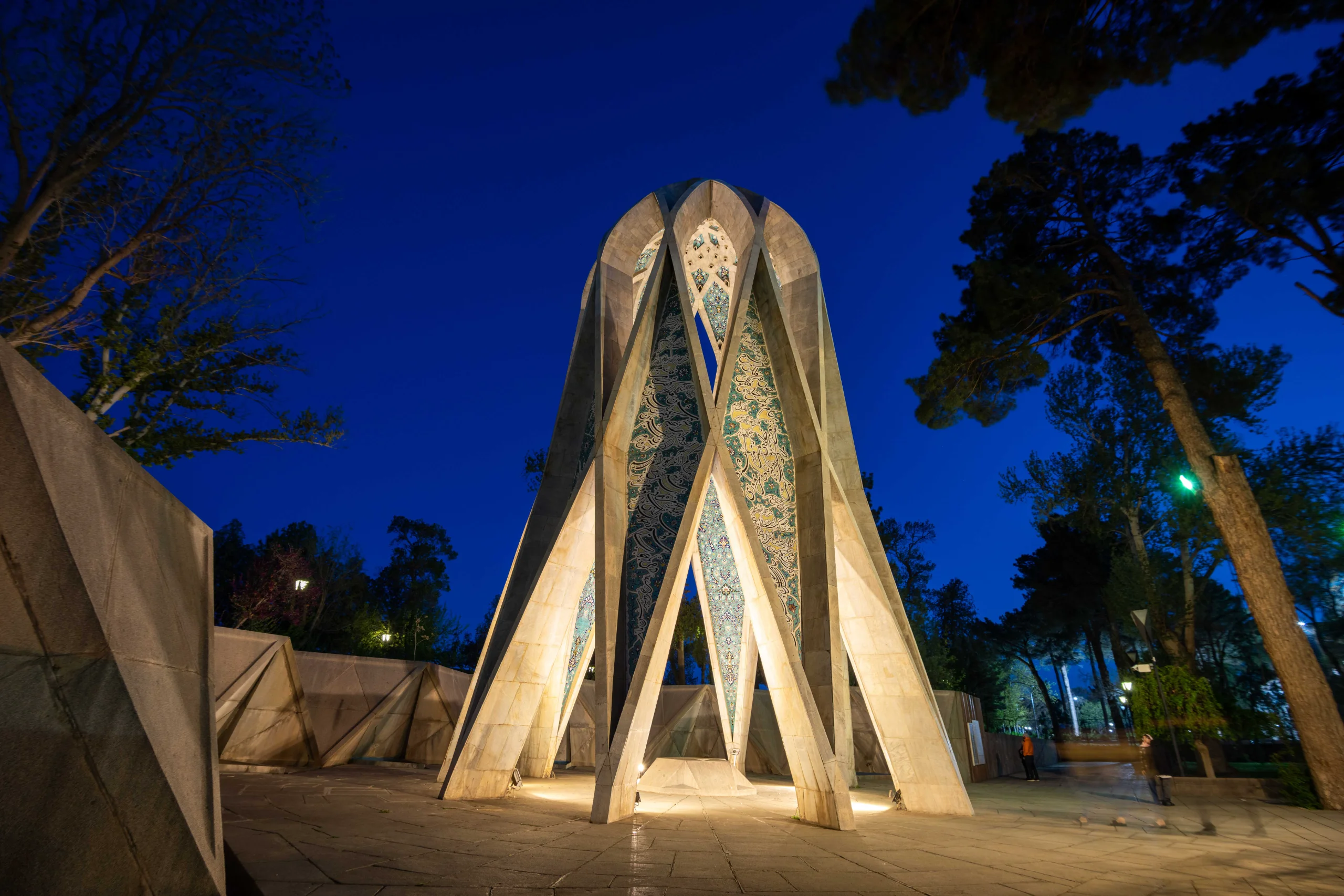 A nighttime wide-angle shot of Khayyam's Mausoleum, a modern conical structure with geometric patterns, illuminated against a deep blue sky, surrounded by dark trees and geometric low walls. Photograph by Mohammad Ataei.