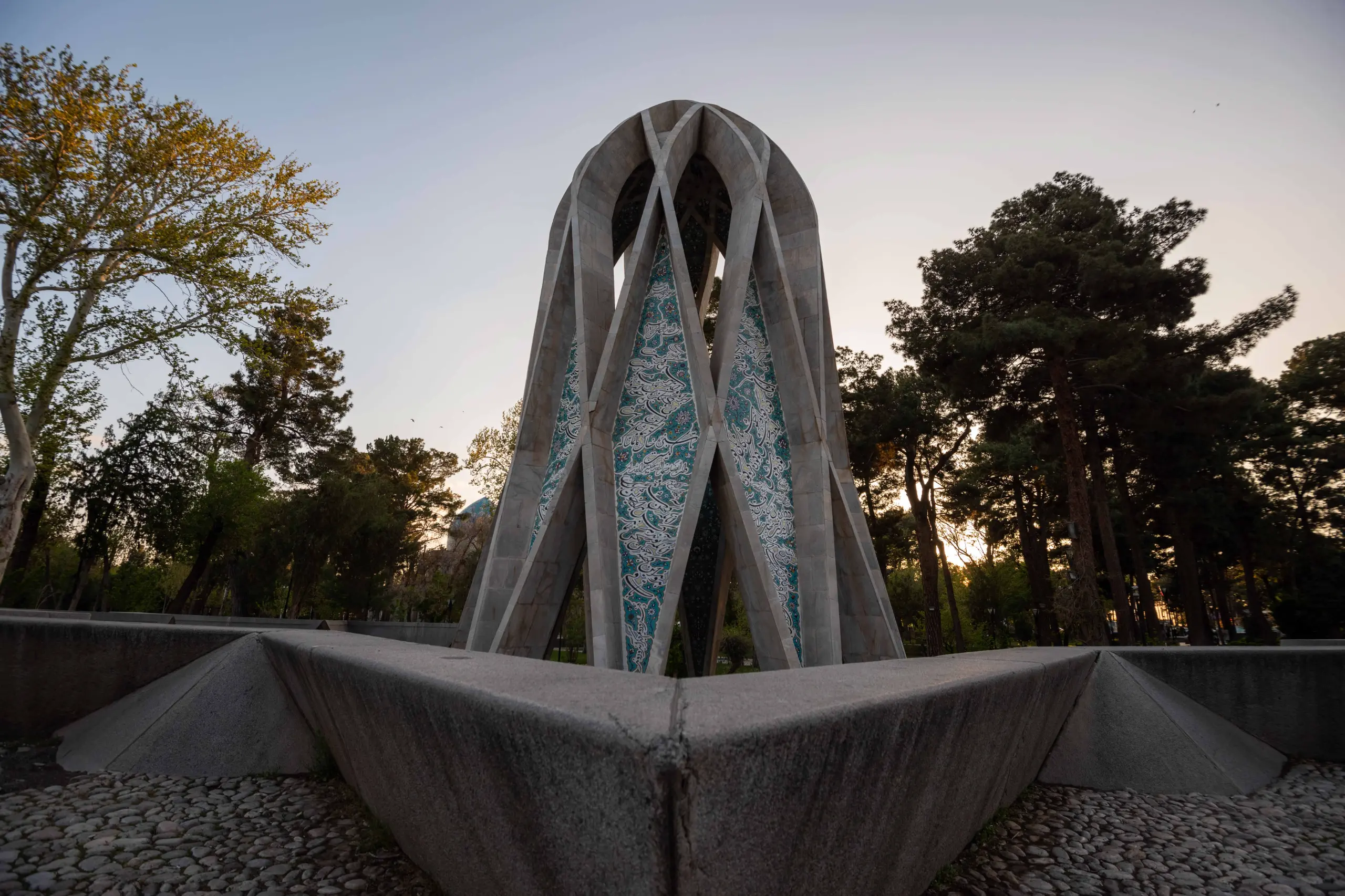 A wide-angle view captures the modern, geometric mausoleum of Omar Khayyam, featuring concrete structures and patterned turquoise tiles, surrounded by trees under a soft, light sky at dusk. Photograph by Mohammad Ataei.