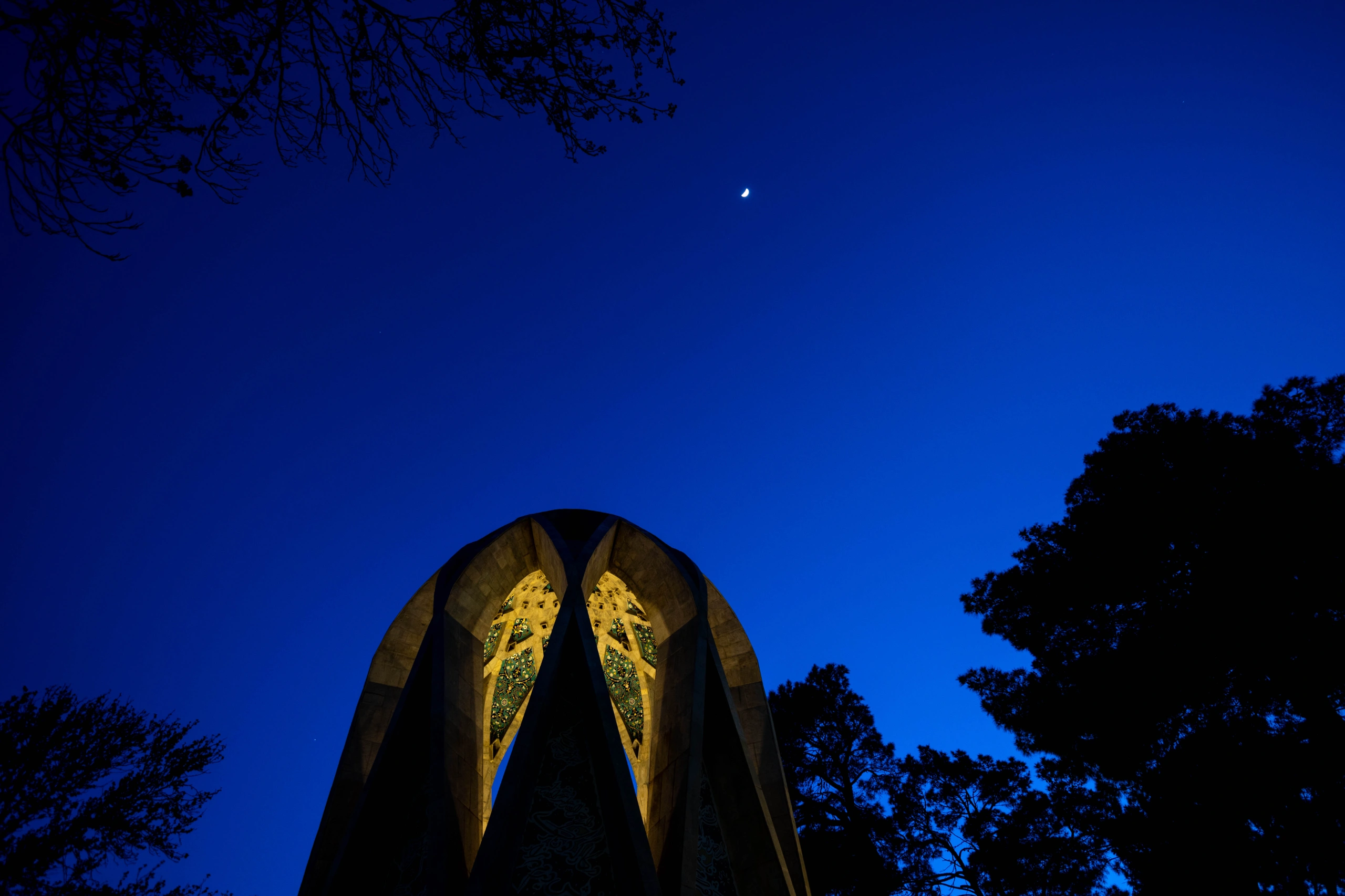 A low-angle shot of the illuminated Khayyam Mausoleum at night, with a crescent moon in a deep blue sky and silhouetted trees. Photograph by Mohammad Ataei.