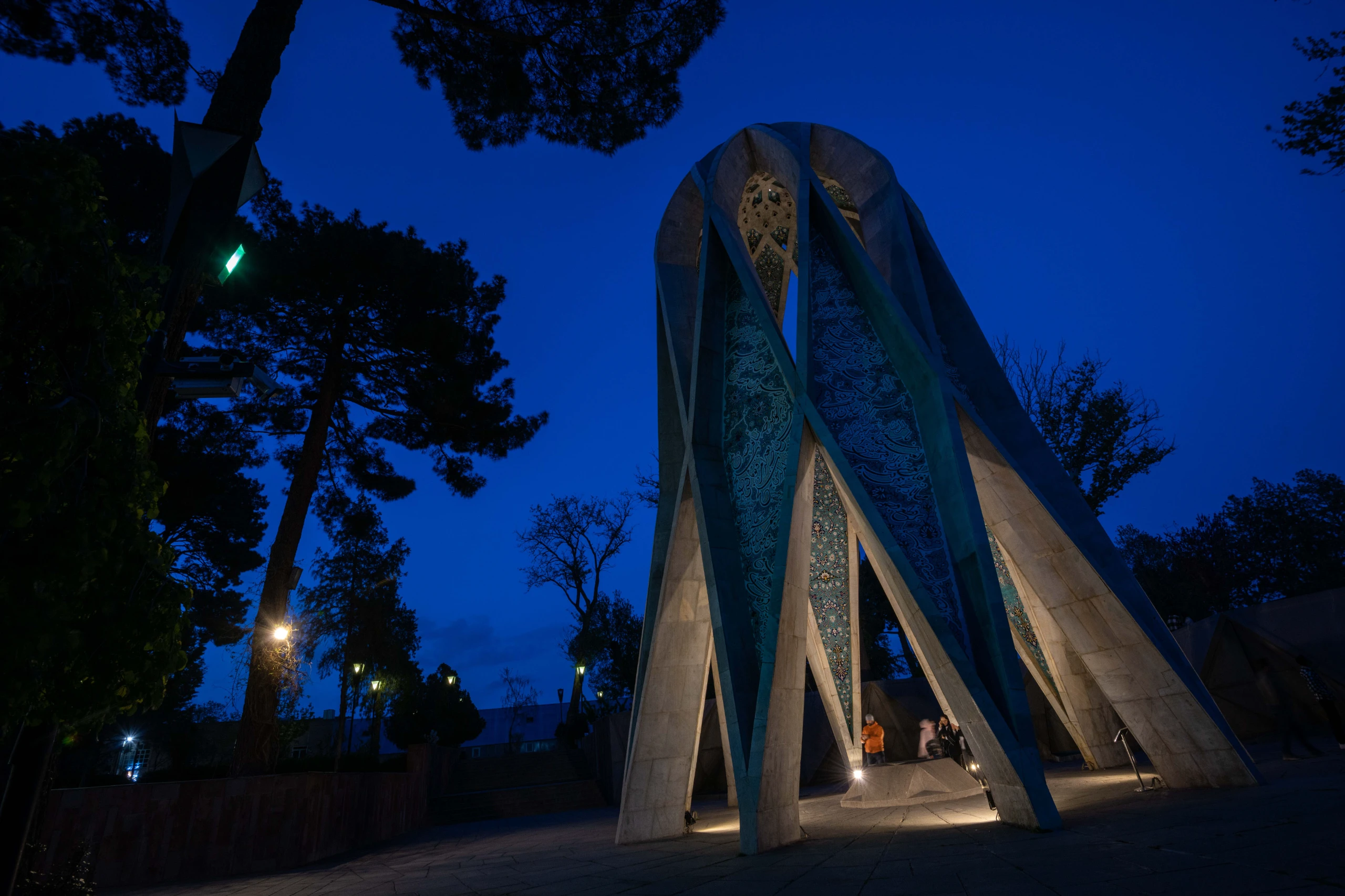 A modern, geometrically designed mausoleum with intricate patterns is illuminated at night against a deep blue sky, surrounded by dark trees. Several people are visible near its base. Photograph by Mohammad Ataei.