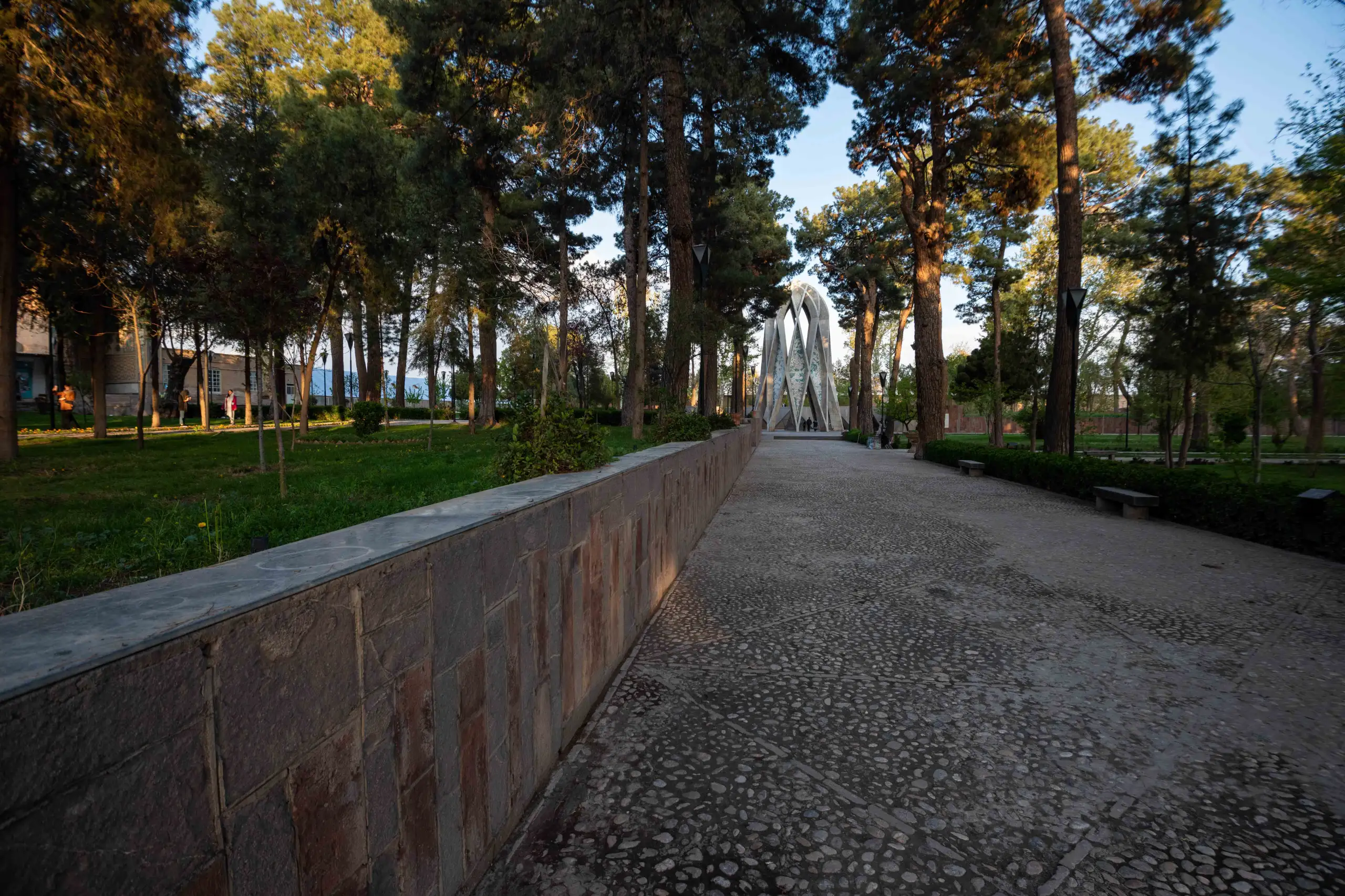 A wide-angle shot of a park pathway leading towards a white, modern, geometric mausoleum structure in the distance, flanked by tall green trees under a clear sky. A stone wall runs along the left side of the pathway, casting shadows from the late afternoon sun. Photograph by Mohammad Ataei.
