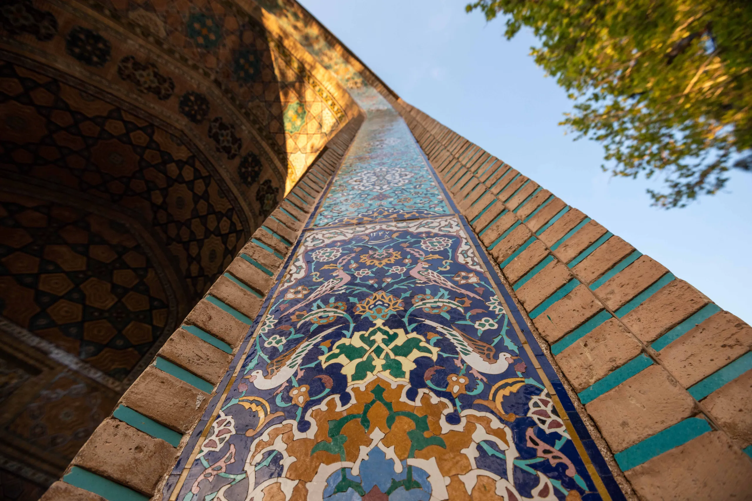 A low-angle shot captures a richly decorated architectural column with blue, green, and gold tiled patterns of birds and floral motifs, next to brickwork, under a clear blue sky. Photograph by Mohammad Ataei.