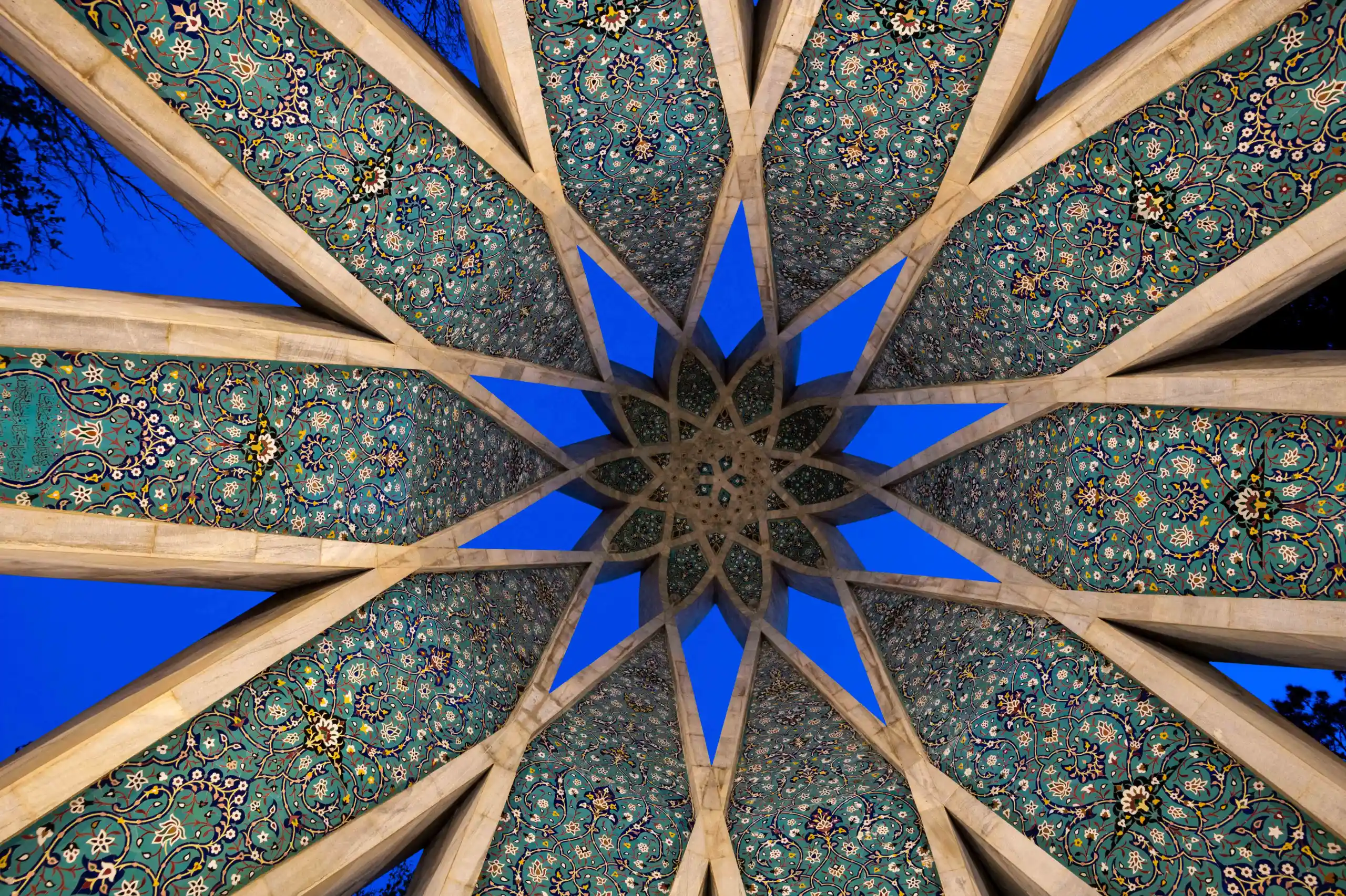 An upward view of a geometrically patterned ceiling with intricate blue, green, and cream tiles forming a star shape, revealing a deep blue sky through its openings. Photograph by Mohammad Ataei.
