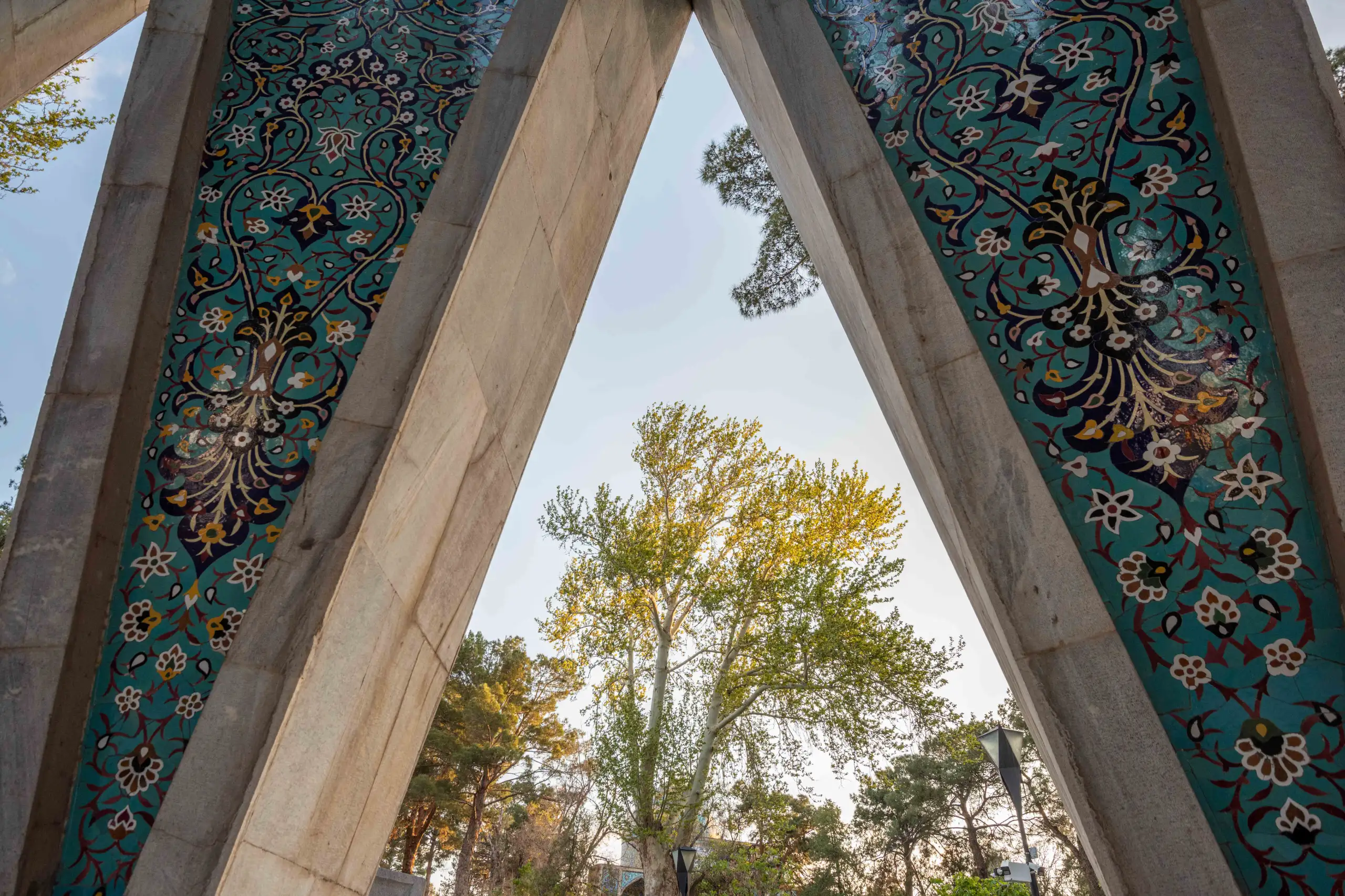 A low-angle view captures the turquoise tilework and concrete structure of Omar Khayyam's Mausoleum, framing a vibrant green tree and sky. Photograph by Mohammad Ataei.