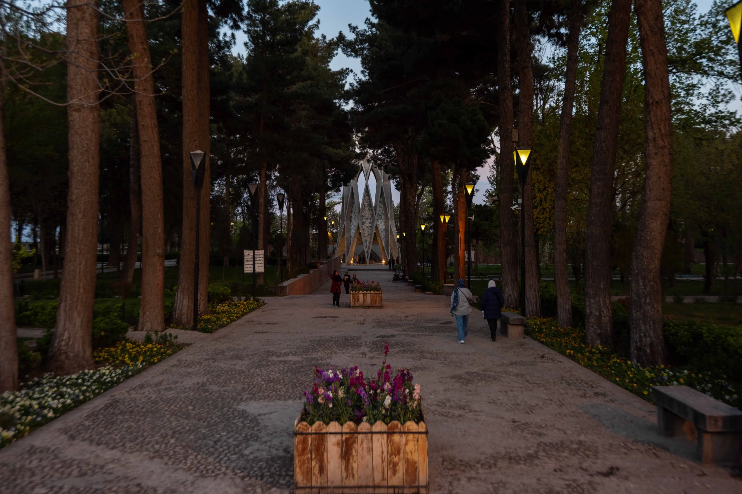 A long, tree-lined path leads to the modern, geometric tomb of Omar Khayyam, illuminated at dusk with people walking on the pathway. Photograph by Mohammad Ataei.
