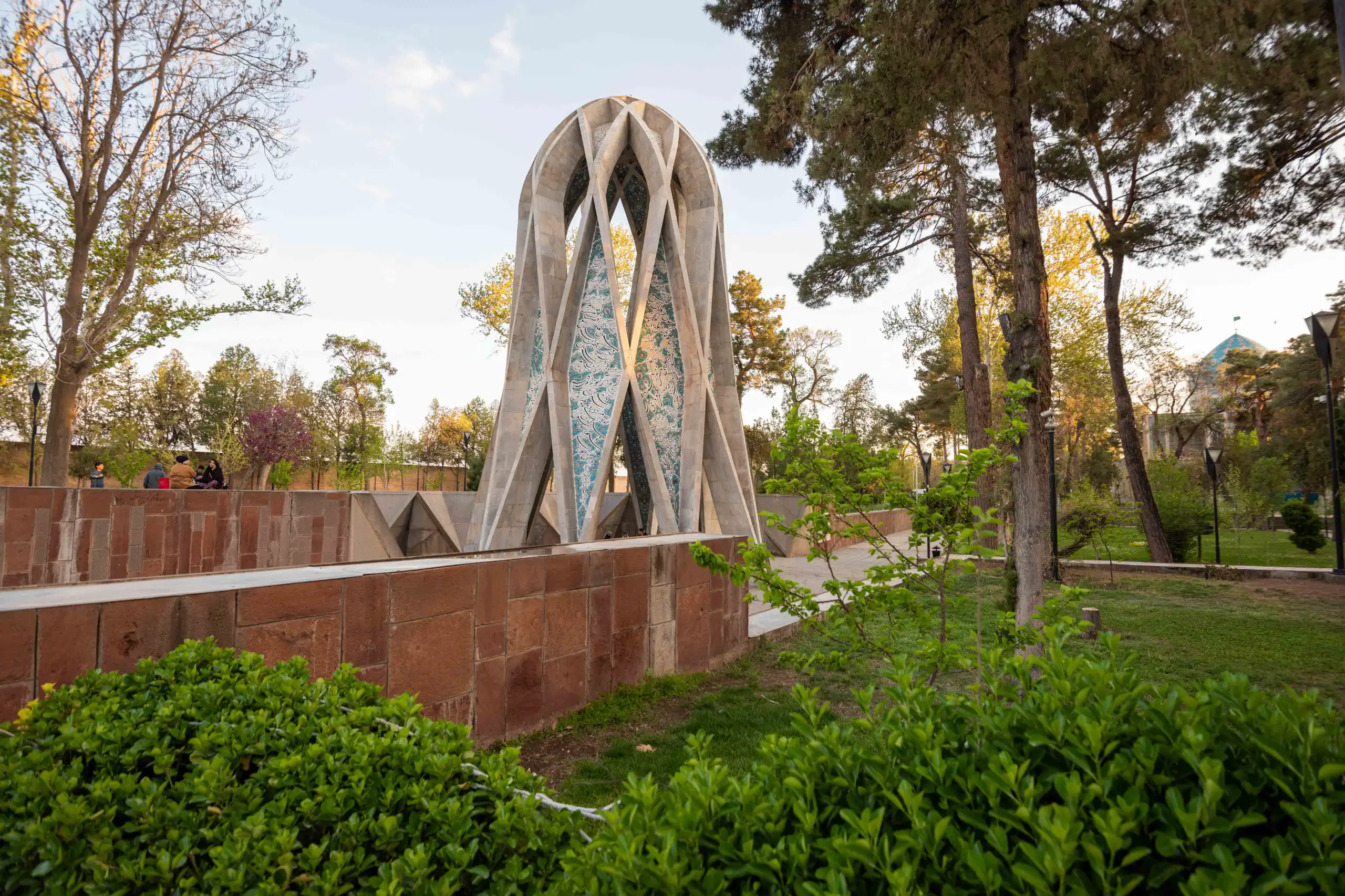 A modern, geometric concrete monument with intricate blue tilework stands prominently in a lush green park, surrounded by tall trees under a bright sky. In the background, people are visible sitting on a low wall, and a blue domed structure can be faintly seen. Photograph by Mohammad Ataei.