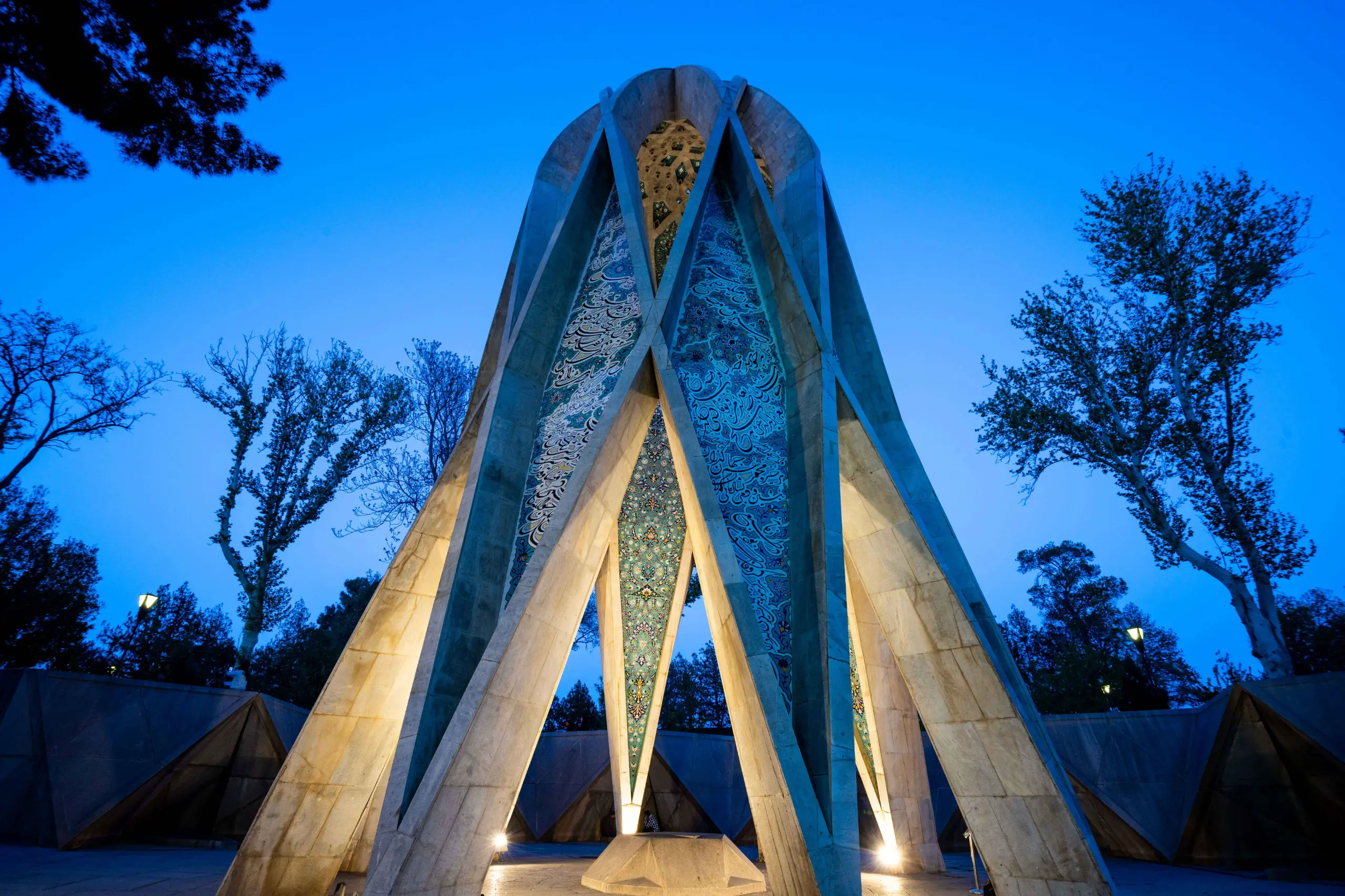 A low-angle shot of Omar Khayyam's Tomb, a towering geometric structure with intricate blue and green tilework, illuminated at twilight against a deep blue sky, surrounded by dark trees. Photograph by Mohammad Ataei.