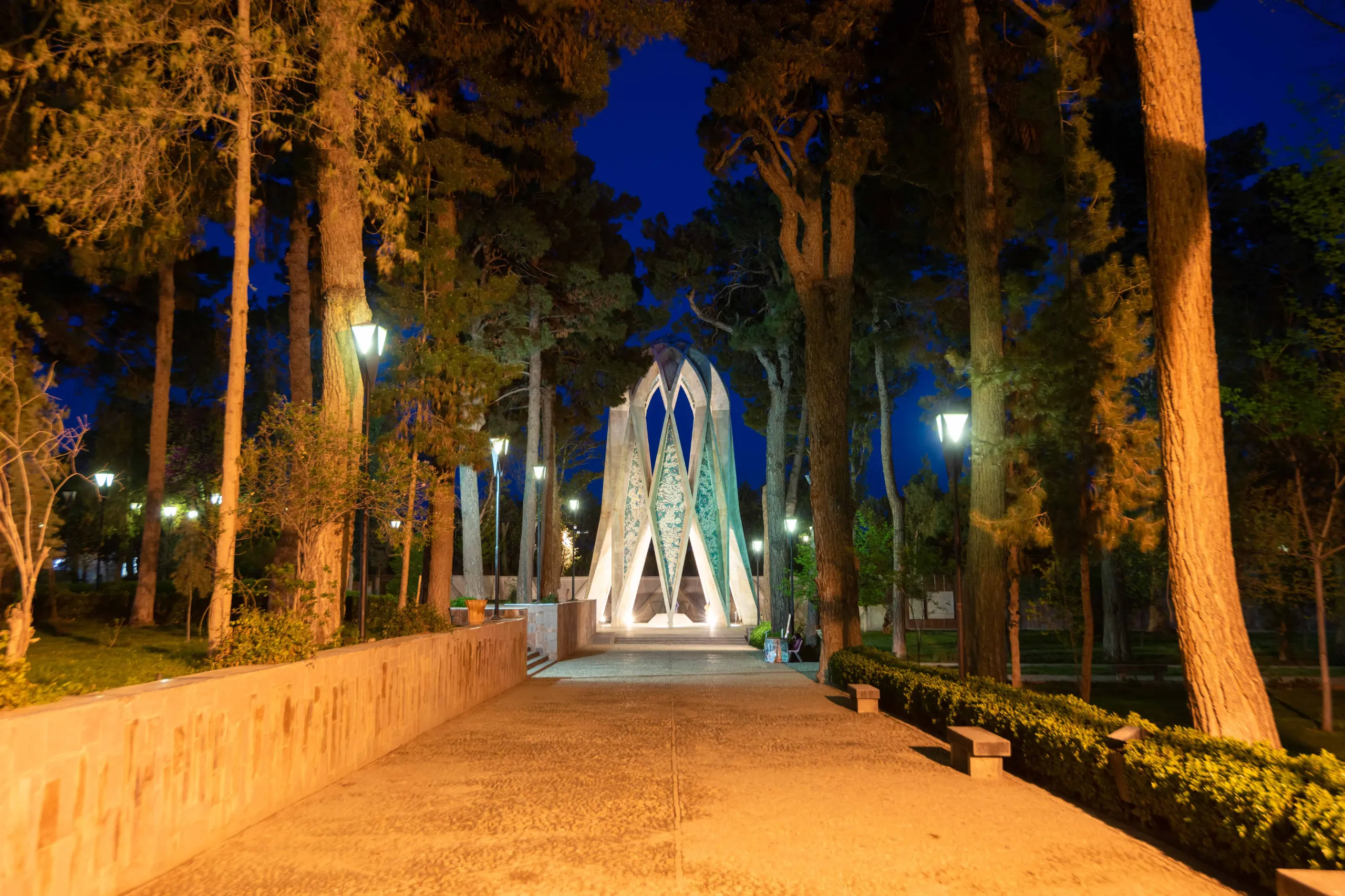 A wide shot of the modern, geometric Tomb of Omar Khayyam, illuminated at night, surrounded by tall trees and a paved pathway. The sky is deep blue. Photograph by Mohammad Ataei.