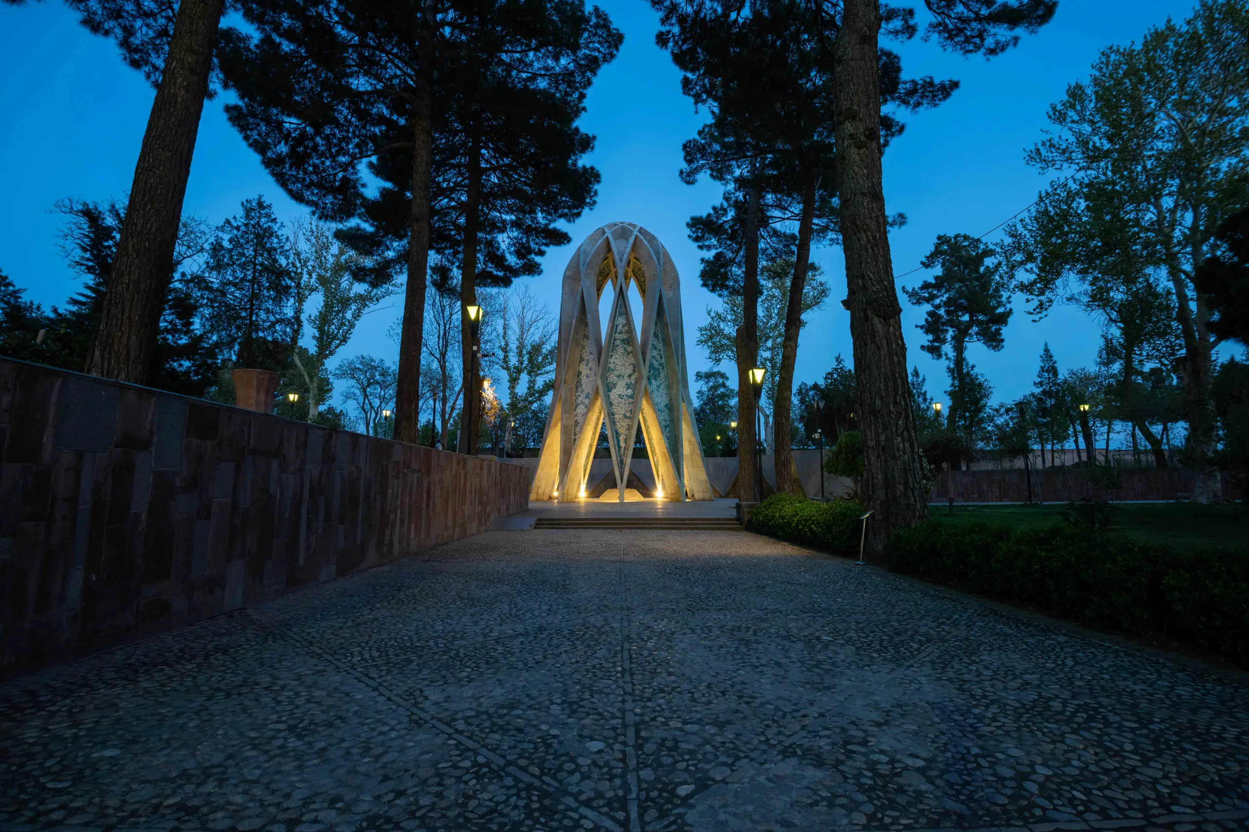 A wide shot of Omar Khayyam's modern, geometric tomb, illuminated from below, standing between tall pine trees at dusk. A paved stone path leads to the monument, with a stone wall on the left. Photograph by Mohammad Ataei.