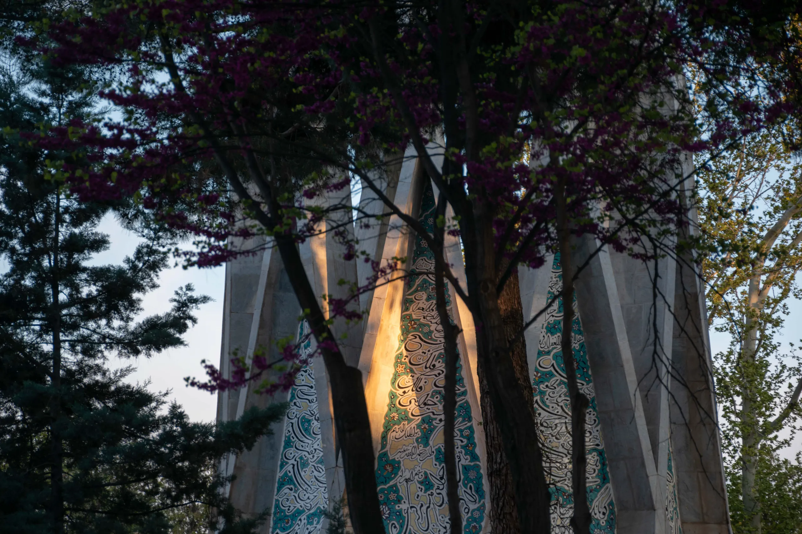 A close-up view of the concrete pillars and turquoise tilework of the Khayyam Mausoleum partially obscured by flowering trees. Photograph by Mohammad Ataei.
