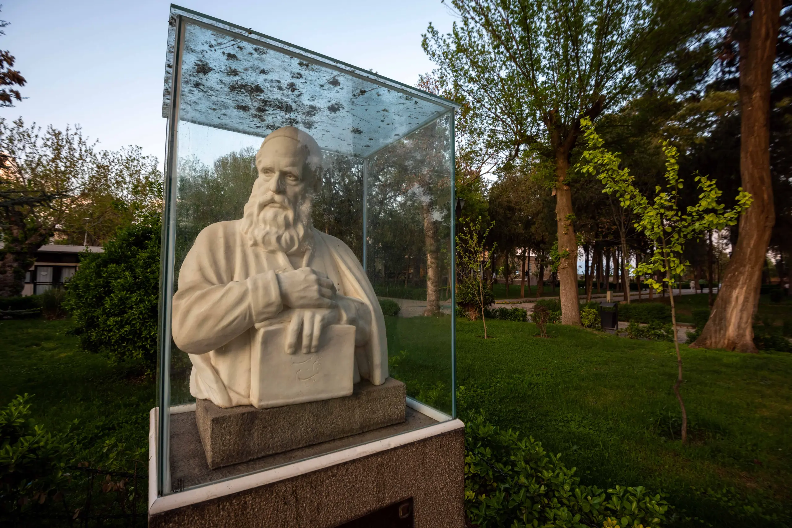 A stone bust of a bearded man holding a book and quill, enclosed in a glass case, set in a green park with trees under a bright sky. Photograph by Mohammad Ataei.