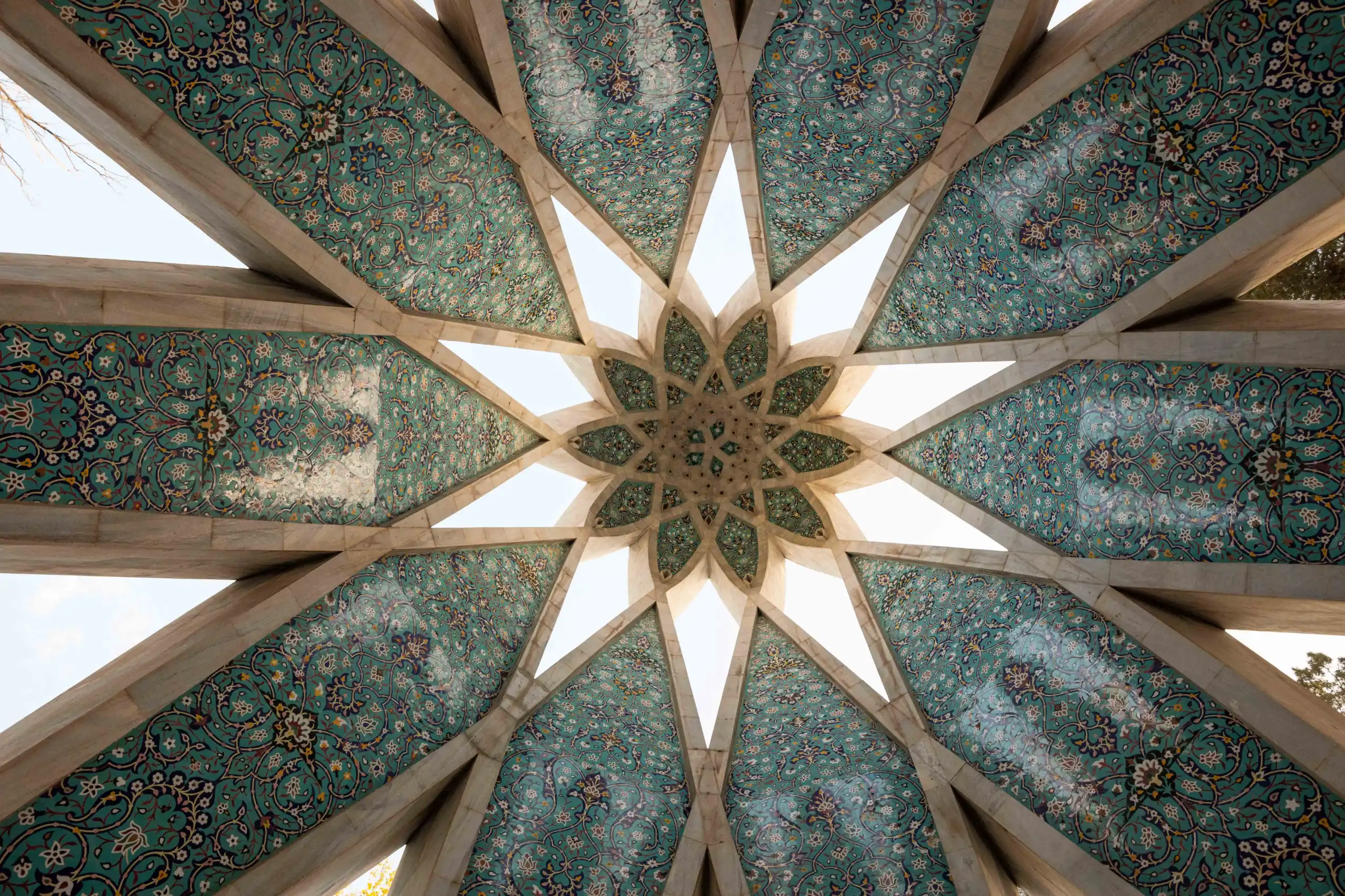 An upward view of an intricately tiled domed ceiling in turquoise and cream, forming a large star pattern with triangular openings allowing glimpses of the sky. Photograph by Mohammad Ataei.