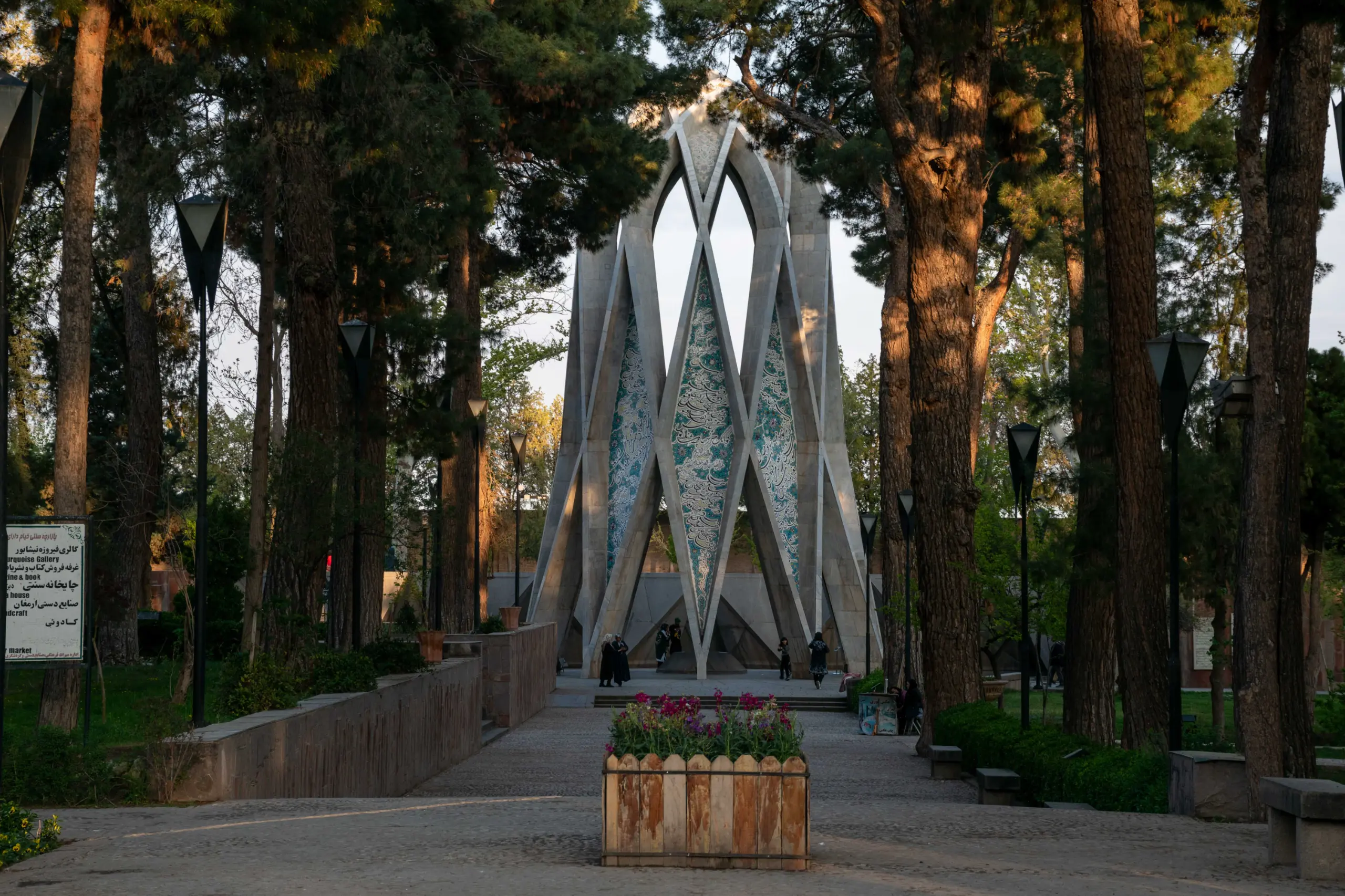 A wide-angle outdoor shot of the Omar Khayyam Mausoleum in Nishapur, featuring its modern geometric structure framed by tall pine trees and a stone pathway under natural daylight. Photograph by Mohammad Ataei.