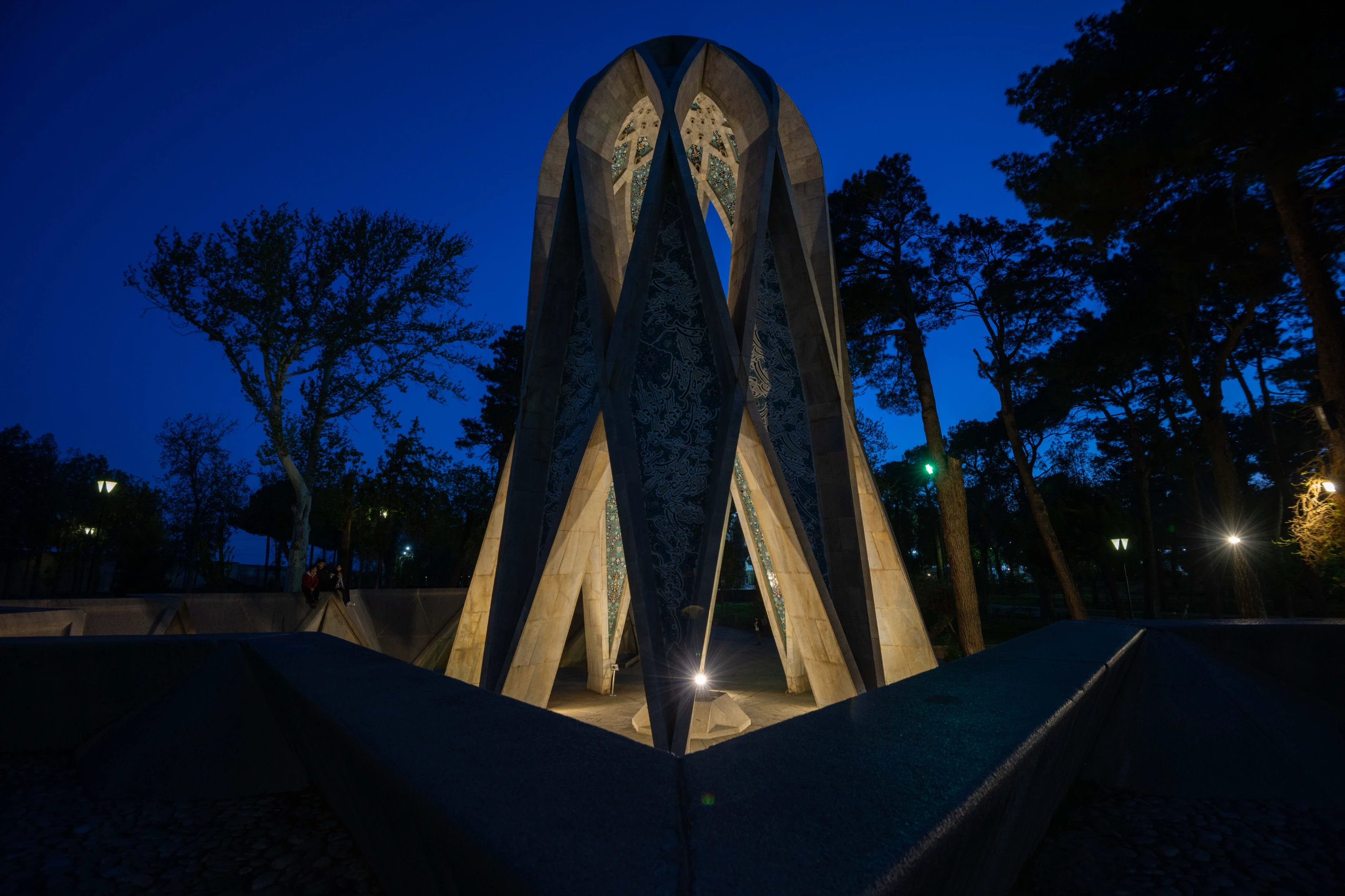A night-time view of the modern, geometric Mausoleum of Omar Khayyam, illuminated against a deep blue sky, surrounded by silhouetted trees. Photograph by Mohammad Ataei.