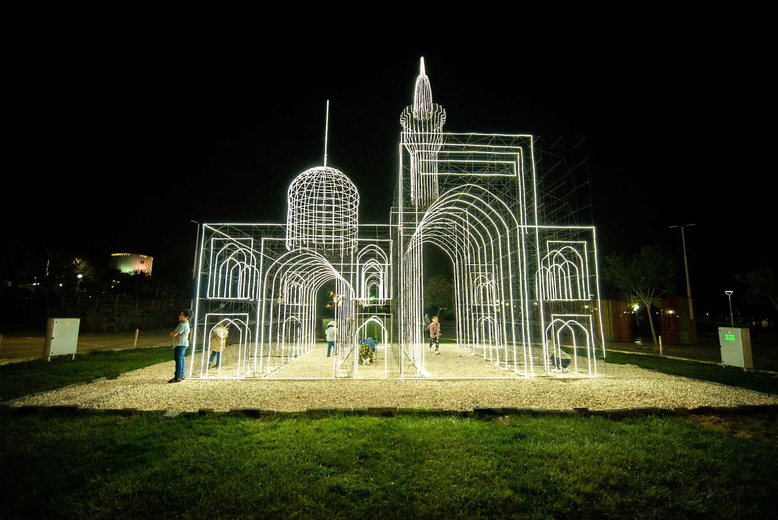 A glowing wireframe light installation depicting an Islamic shrine stands in a park at night, with several children playing nearby. Photograph by Mohammad Ataei.