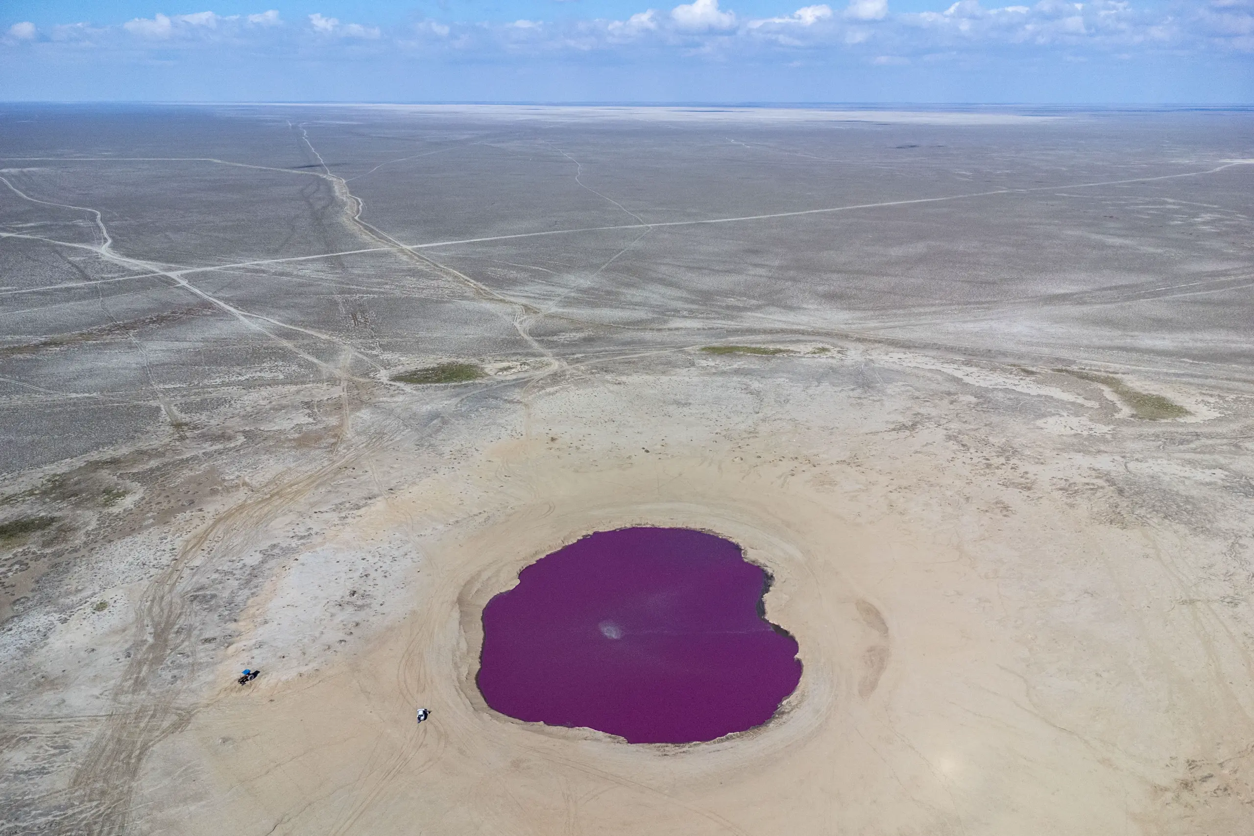 An aerial shot shows a vibrant purple mud volcano pool situated in the middle of a flat, arid landscape under a blue sky with scattered clouds. Photograph by Mohammad Ataei.