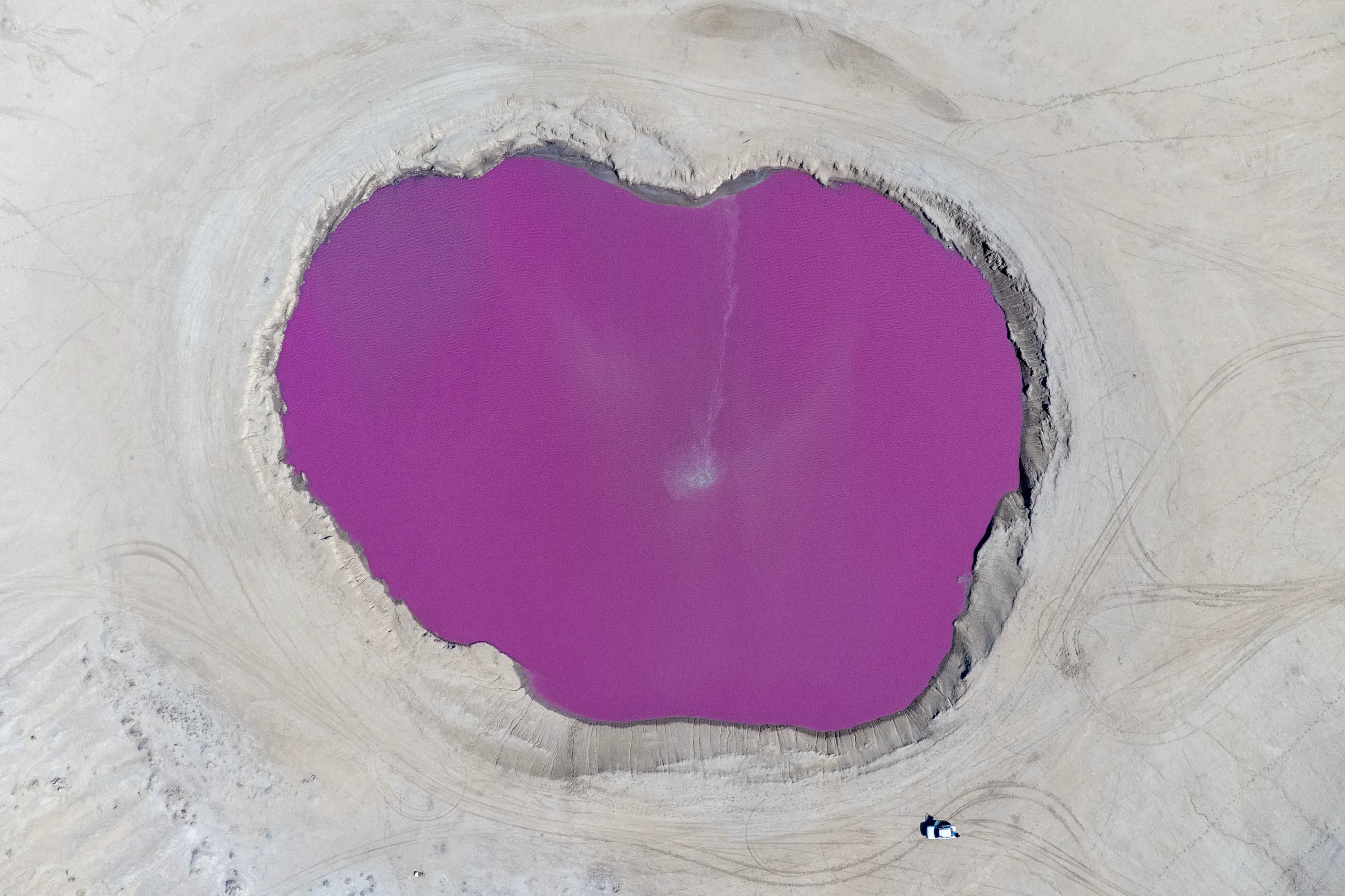 An overhead aerial shot captures a vibrant pink and purple pool inside the crater of the Naftlijeh Mud Volcano surrounded by flat, light-colored terrain. Photograph by Mohammad Ataei.