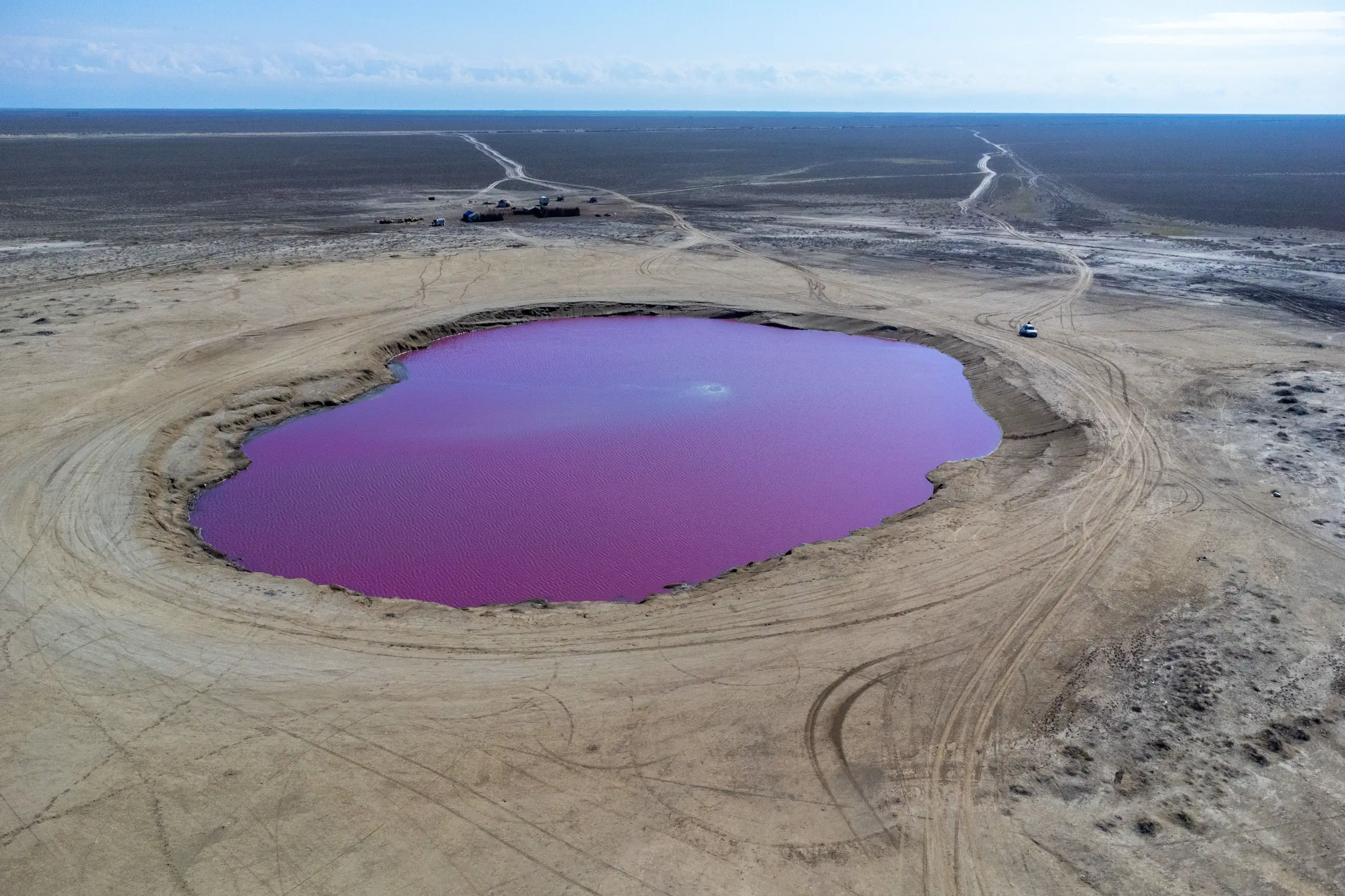 Aerial view of a vibrant pink and purple mud volcano surrounded by flat arid terrain and vehicle tracks, under an open horizon. Photograph by Mohammad Ataei.