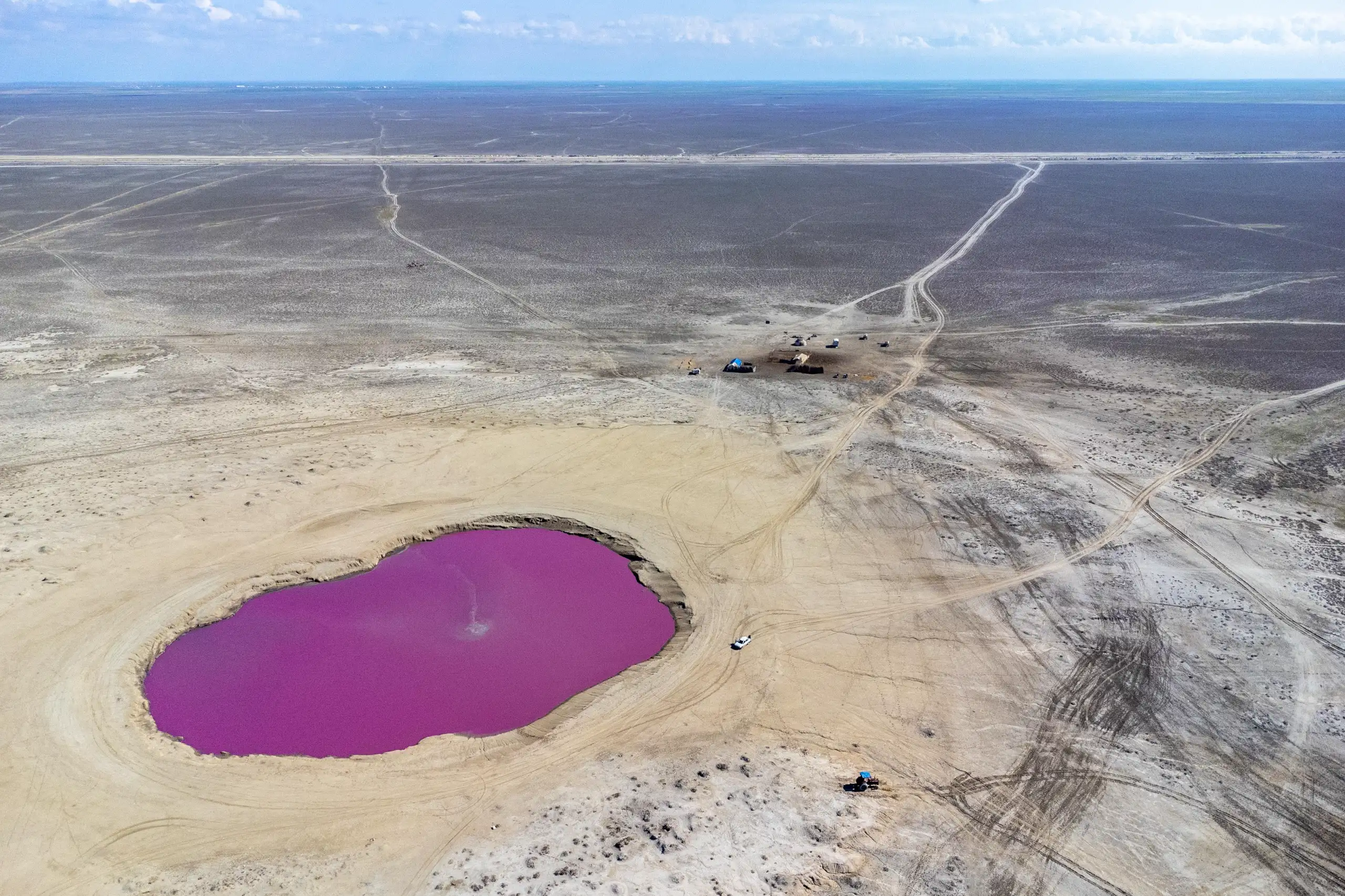 An aerial view shows a vibrant pink mud volcano pool set within a vast, flat arid landscape under a blue sky, with distant tracks and a small settlement visible. Photograph by Mohammad Ataei.
