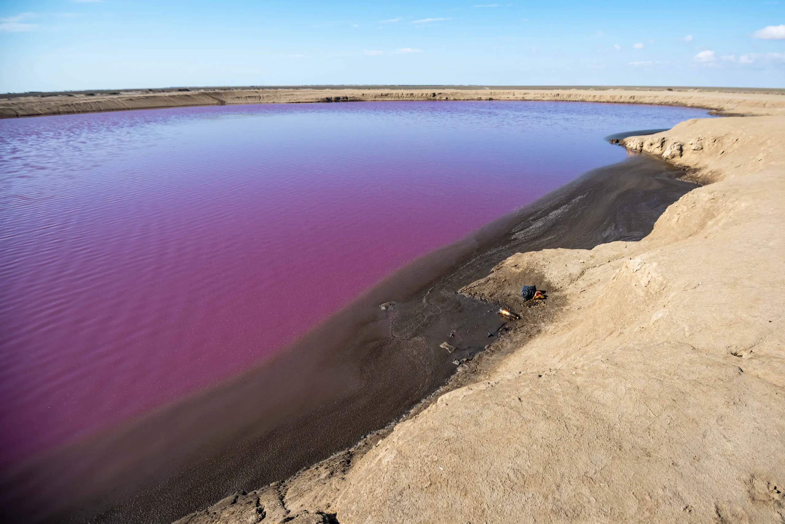 High-angle view of the vibrant pink and purple pool of Naftlijeh Mud Volcano surrounded by sandy banks and dark mud under a blue sky. Photograph by Mohammad Ataei.