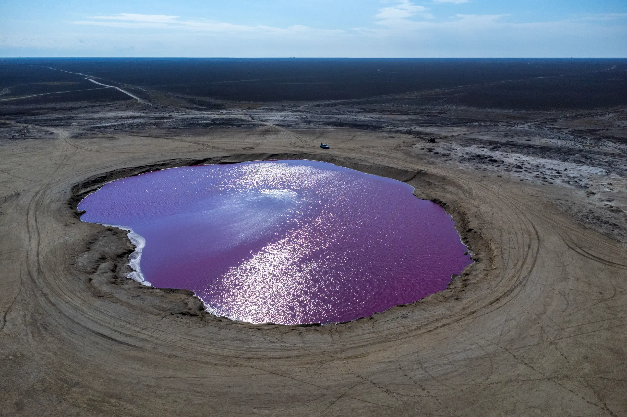 Aerial view of a vibrant pink and purple mud volcano pool surrounded by barren flat terrain and tire tracks under a bright sky. Photograph by Mohammad Ataei.