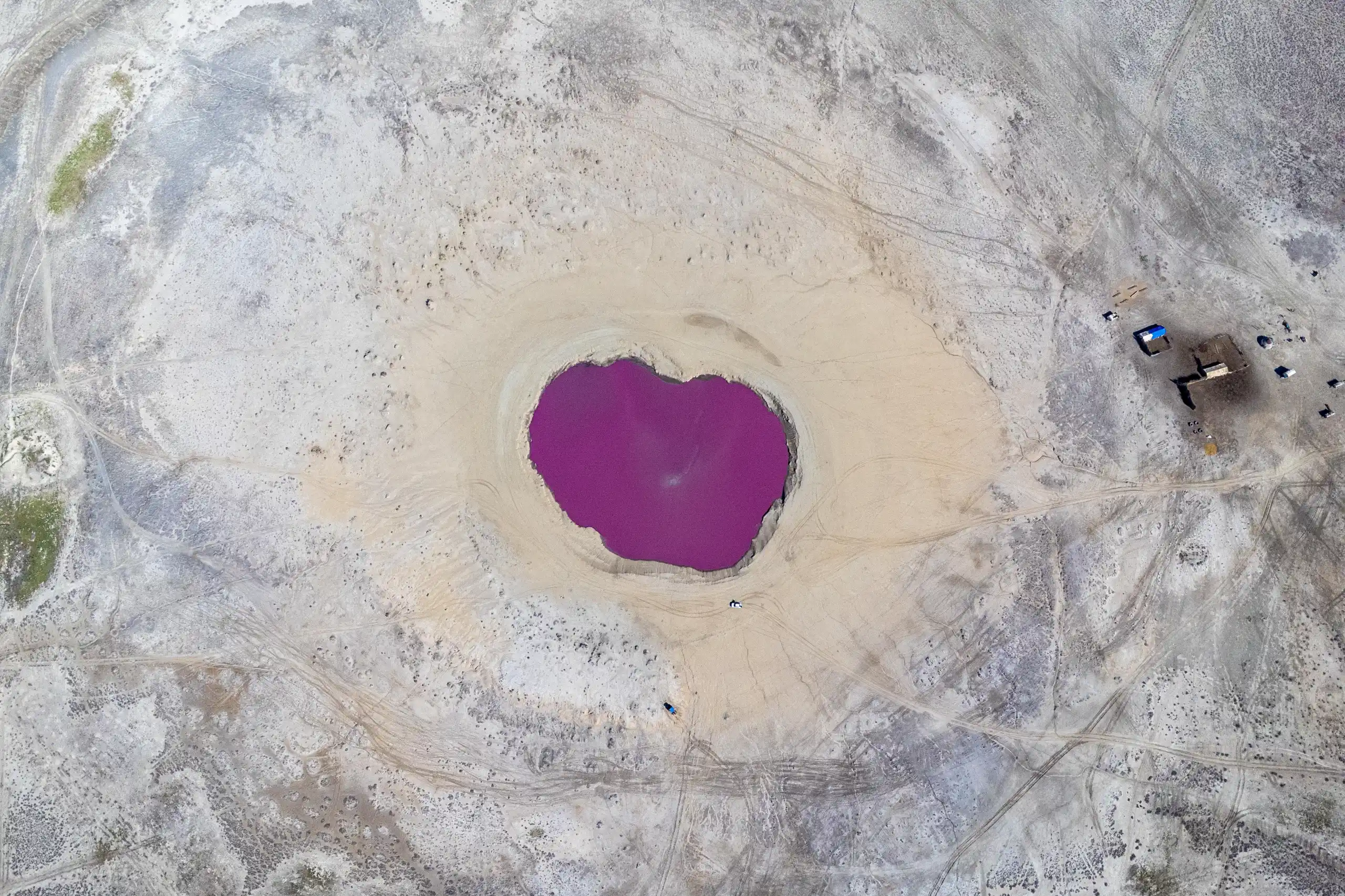 An aerial view captures a vibrant purple mud volcano pool surrounded by pale, dry earth and vehicle tracks. Photograph by Mohammad Ataei.