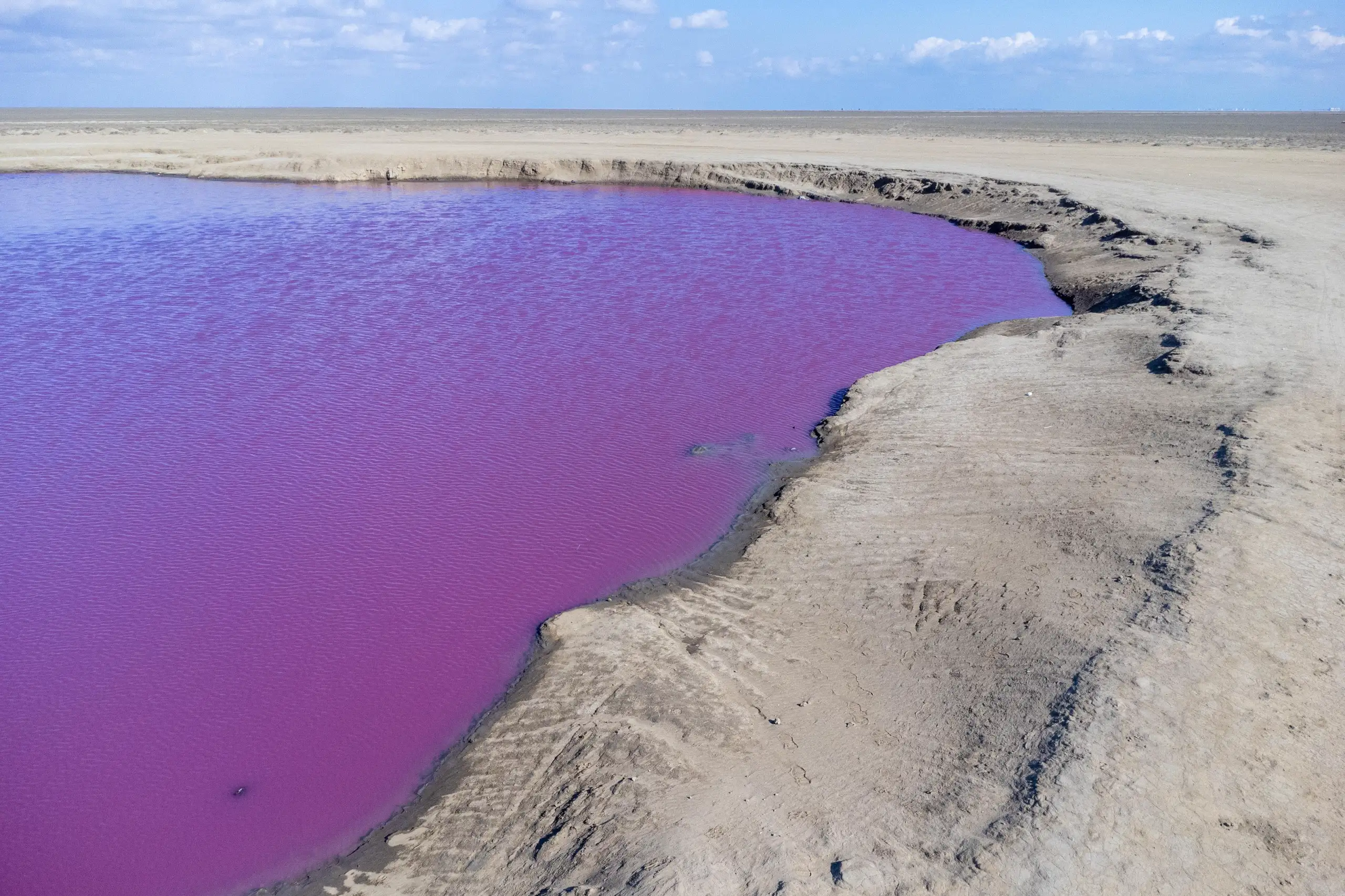 A high-angle view shows a vibrant pink and purple mud volcano pool surrounded by dry, sandy earth under a blue sky with light clouds. Photograph by Mohammad Ataei.