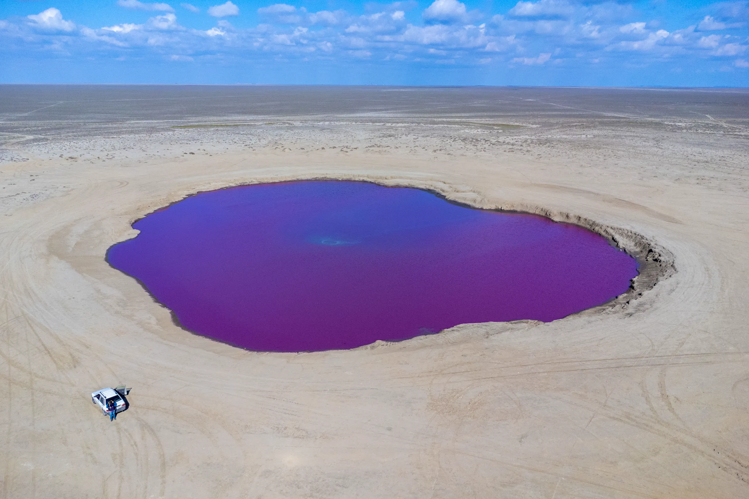 An aerial view shows the vibrant purple-pink basin of the Naftlijeh Mud Volcano surrounded by flat desert terrain, with a small white car parked nearby under a partly cloudy sky. Photograph by Mohammad Ataei.