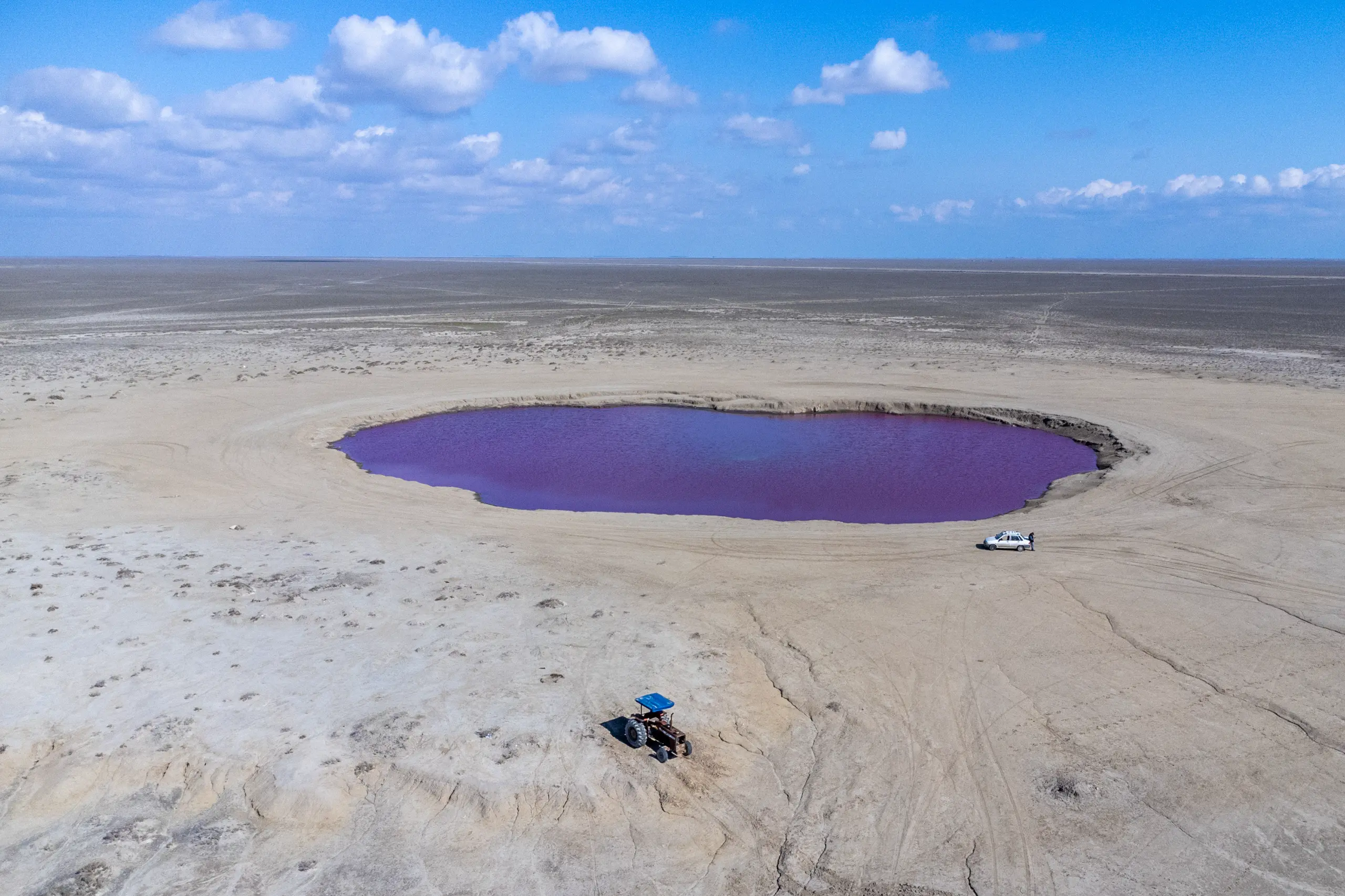 Aerial view of a striking purple-pink mud volcano pool in a vast arid coastal plain, with a tractor and a small car parked nearby under a cloudy blue sky. Photograph by Mohammad Ataei.