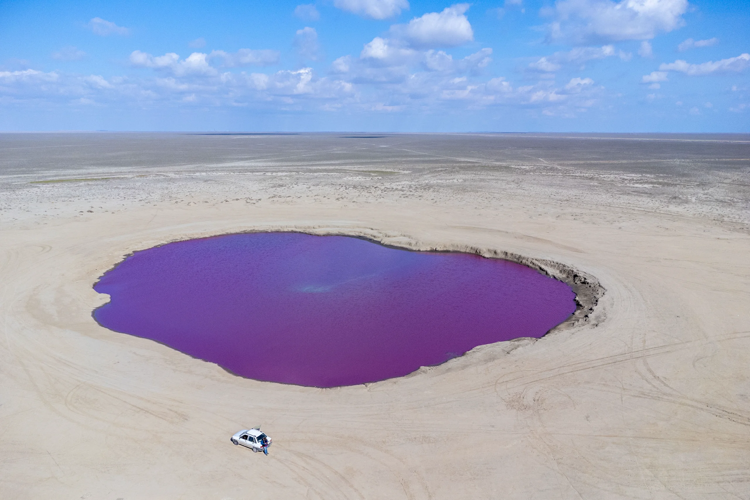 An aerial view shows a vibrant purple mud volcano crater pool surrounded by flat sandy terrain under a blue sky with scattered clouds, with a small car parked nearby. Photograph by Mohammad Ataei.