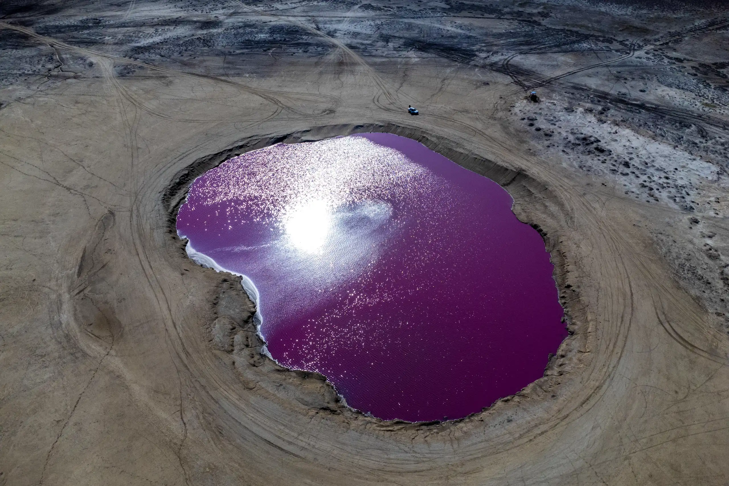 An aerial photograph looking straight down at the vibrant purple and pink pool of the Naftlijeh Mud Volcano surrounded by dry desert-like terrain and tire tracks. Photograph by Mohammad Ataei.