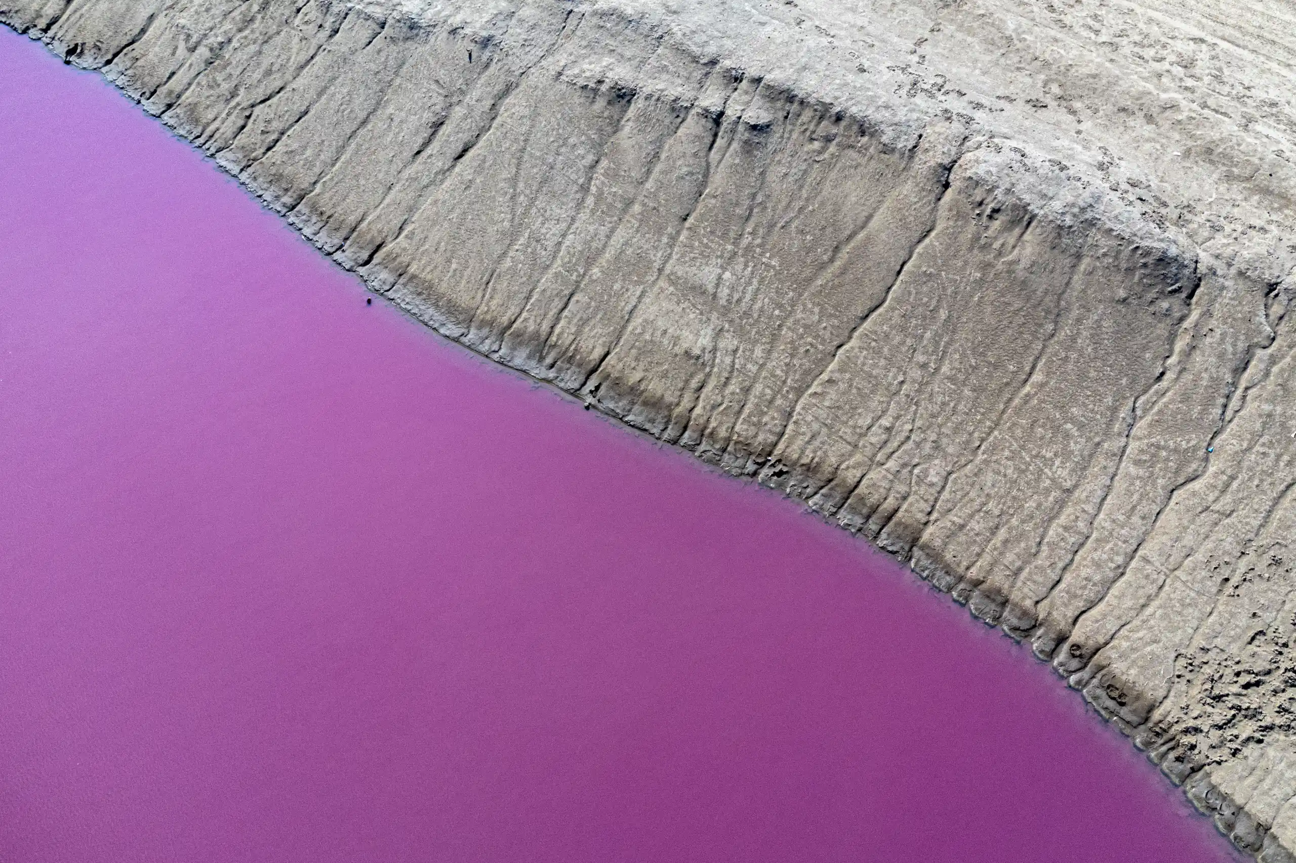 An aerial view showing a vibrant purple-pink pool of water alongside an eroded, textured soil bank of a mud volcano. Photograph by Mohammad Ataei.