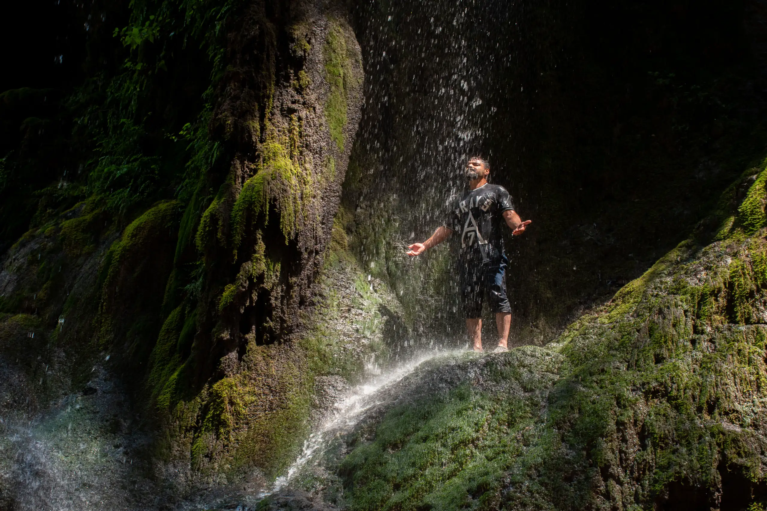 A man standing beneath the cascading water of a mossy waterfall with arms outstretched in Gorgan, Golestan Province, Iran. Photograph by Mohammad Ataei.