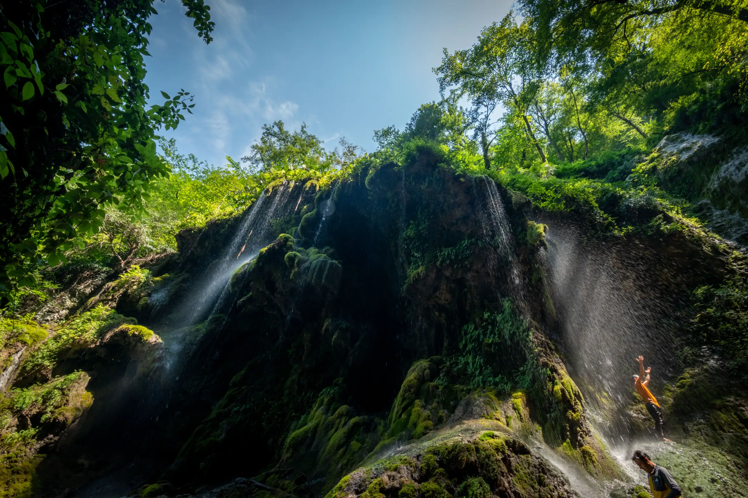A wide-angle view of Behesht Baran Waterfall cascading down mossy rock formations surrounded by dense green trees in Gorgan, with two people standing near the falling water. Photograph by Mohammad Ataei.