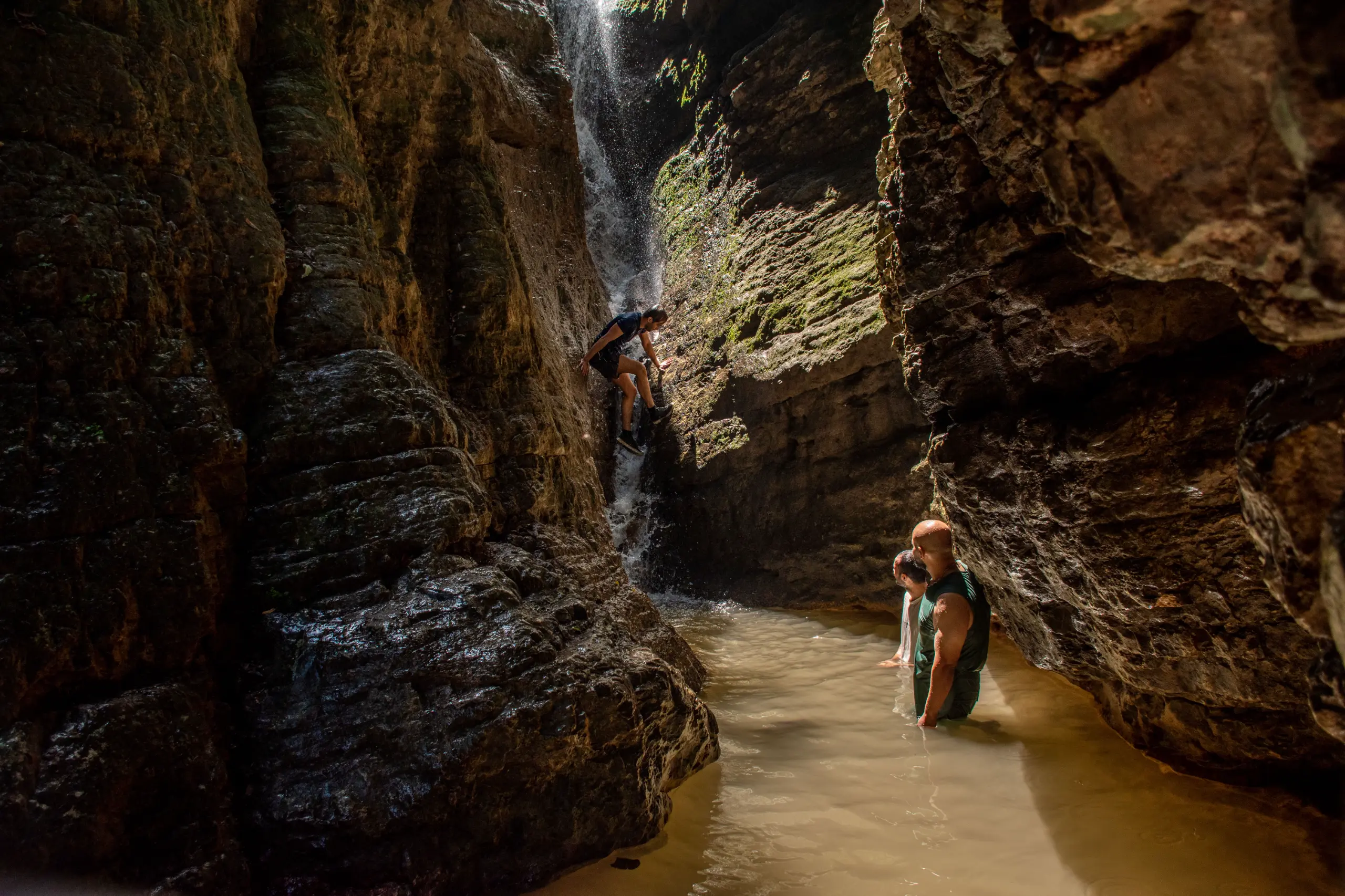 Travelers wading through water in a narrow rocky canyon while another person climbs a small waterfall in Gorgan, Iran. Photograph by Mohammad Ataei.