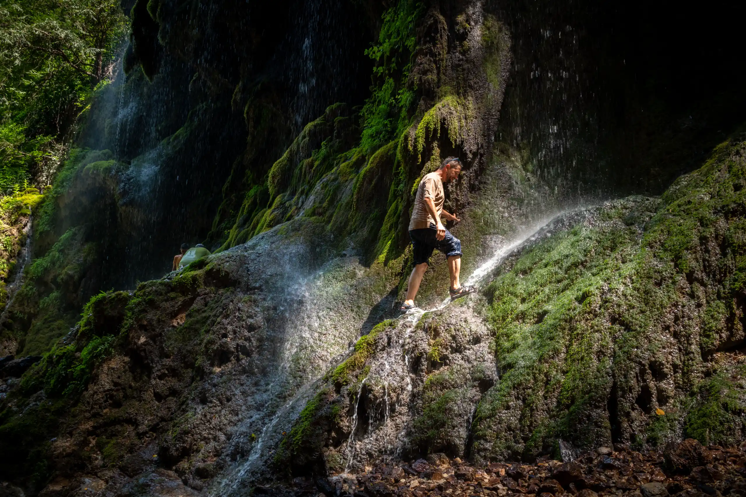 A man in a light shirt walks on a mossy, wet rock wall beside a cascading waterfall, with lush green foliage surrounding the cliff. Photograph by Mohammad Ataei.