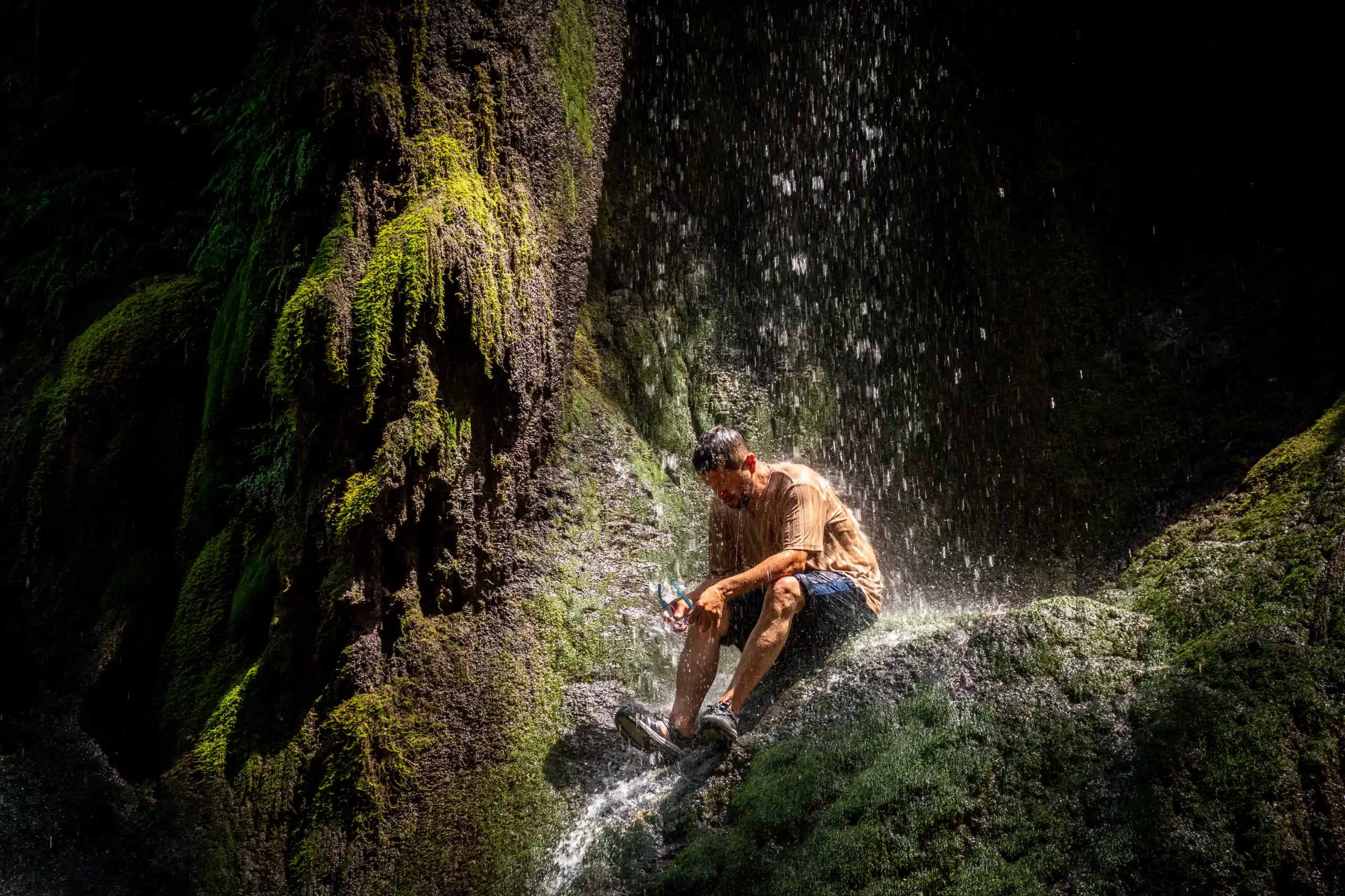 A man sits on a mossy rock underneath a cascading waterfall, getting wet from the falling water. Photograph by Mohammad Ataei.