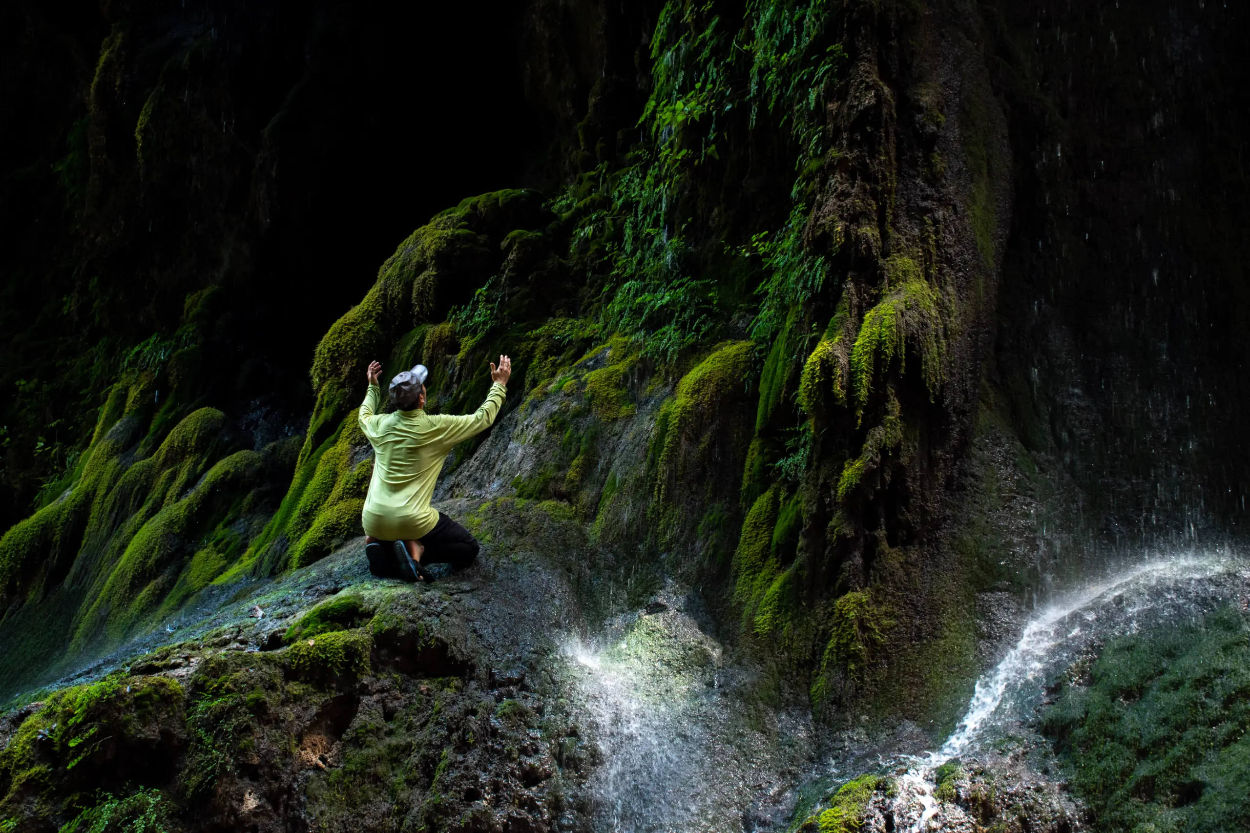 A traveler in a light shirt kneels on a mossy rock with raised hands inside the misty, moss-covered grotto of Behesht Baran Waterfall in Gorgan, Iran. Photograph by Mohammad Ataei.
